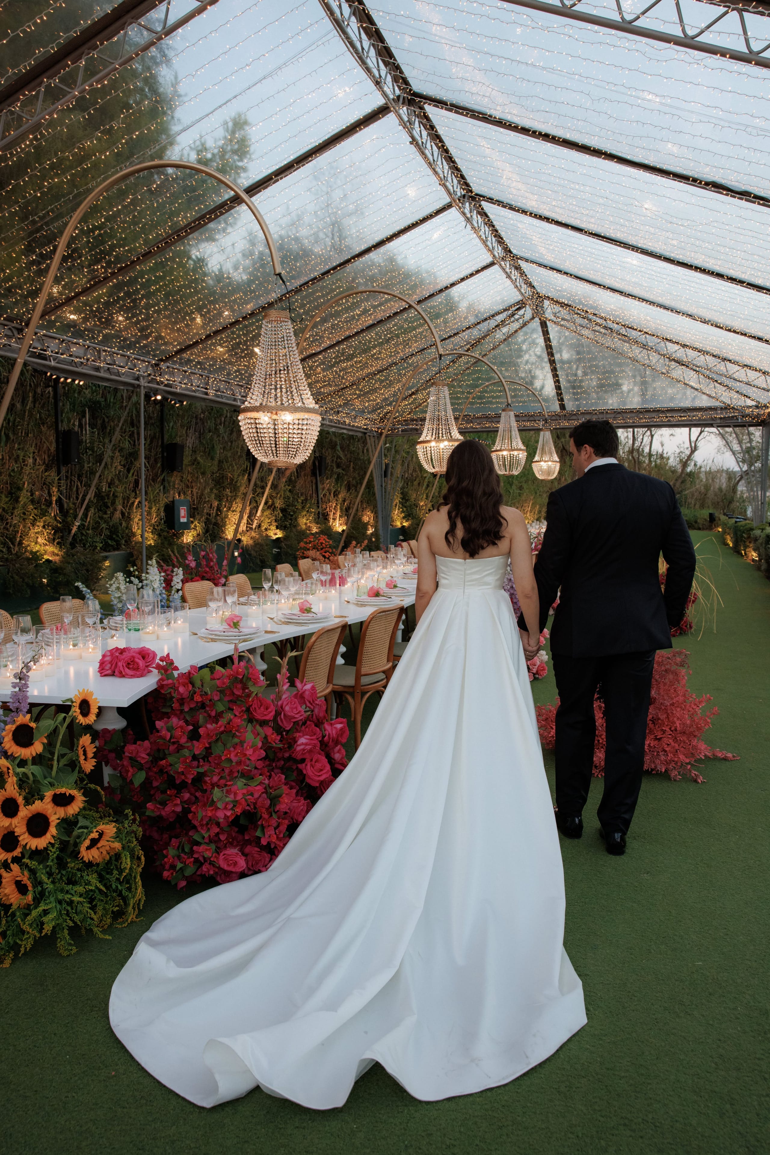 A bride in a strapless satin gown with a long sweeping train walks hand in hand with her groom in a black suit, seen from behind, along a green lawn beneath a clear canopy strung with fairy lights. Beside them a long white table is set for a wedding dinner at Island Club & Restaurant in Vari, Athens, Greece, its low floral displays of fuchsia bougainvillea, pink roses and sunflowers glowing in the warm evening light.