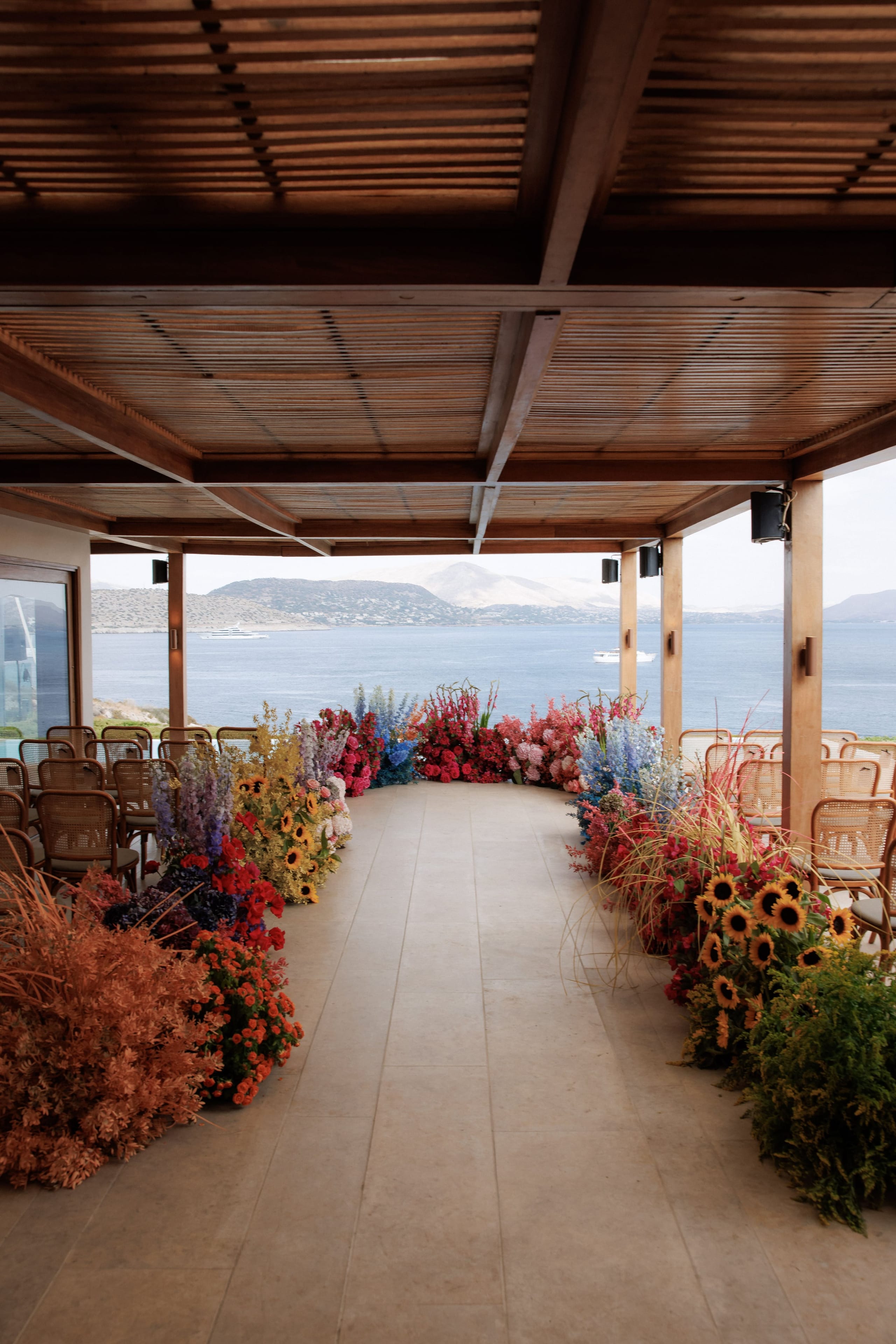 A long aisle on a covered terrace at Island Club & Restaurant in Vari, Athens, Greece, lined on both sides with wedding flowers in bold reds, oranges, and violet-blue. Tall delphinium spires, sunflowers, and feathery grasses rise from the floor, while cane chairs wait under a slatted wooden pergola. Beyond the open sides, a calm blue sea and hazy hills stretch into the distance under soft, late-afternoon light.