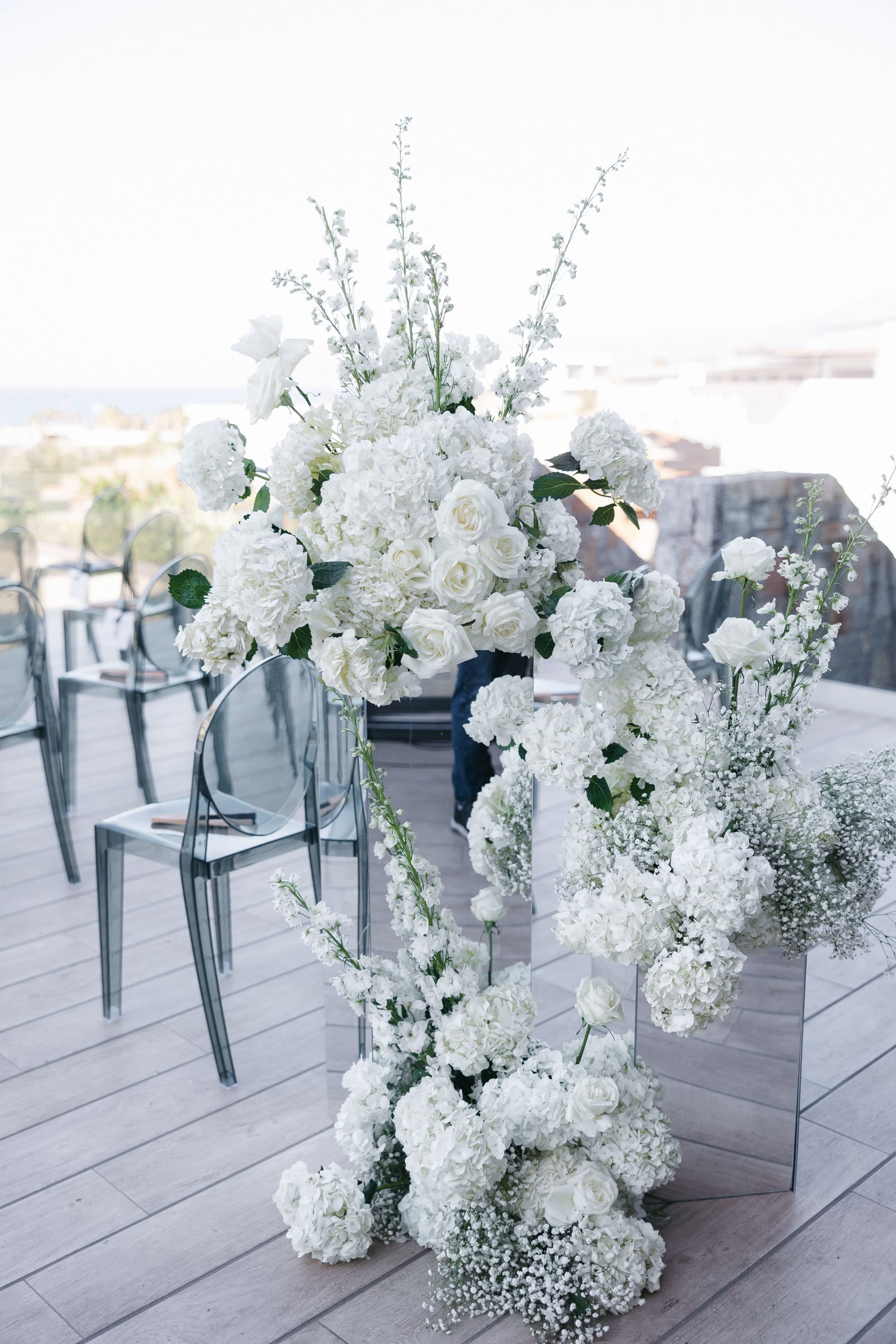 Tall all-white ceremony arrangements of hydrangeas, white garden roses, delphinium spires and baby's breath rise from tall mirrored pedestals on a pale wooden terrace at Abaton Island Resort & Spa on Crete, Greece. Clear acrylic Ghost chairs sit in rows behind them, with a soft blue sea and hazy hillside stretching into the bright, misty distance. The overall feel is airy, elegant and entirely monochrome, a wedding aisle styled in pure white.