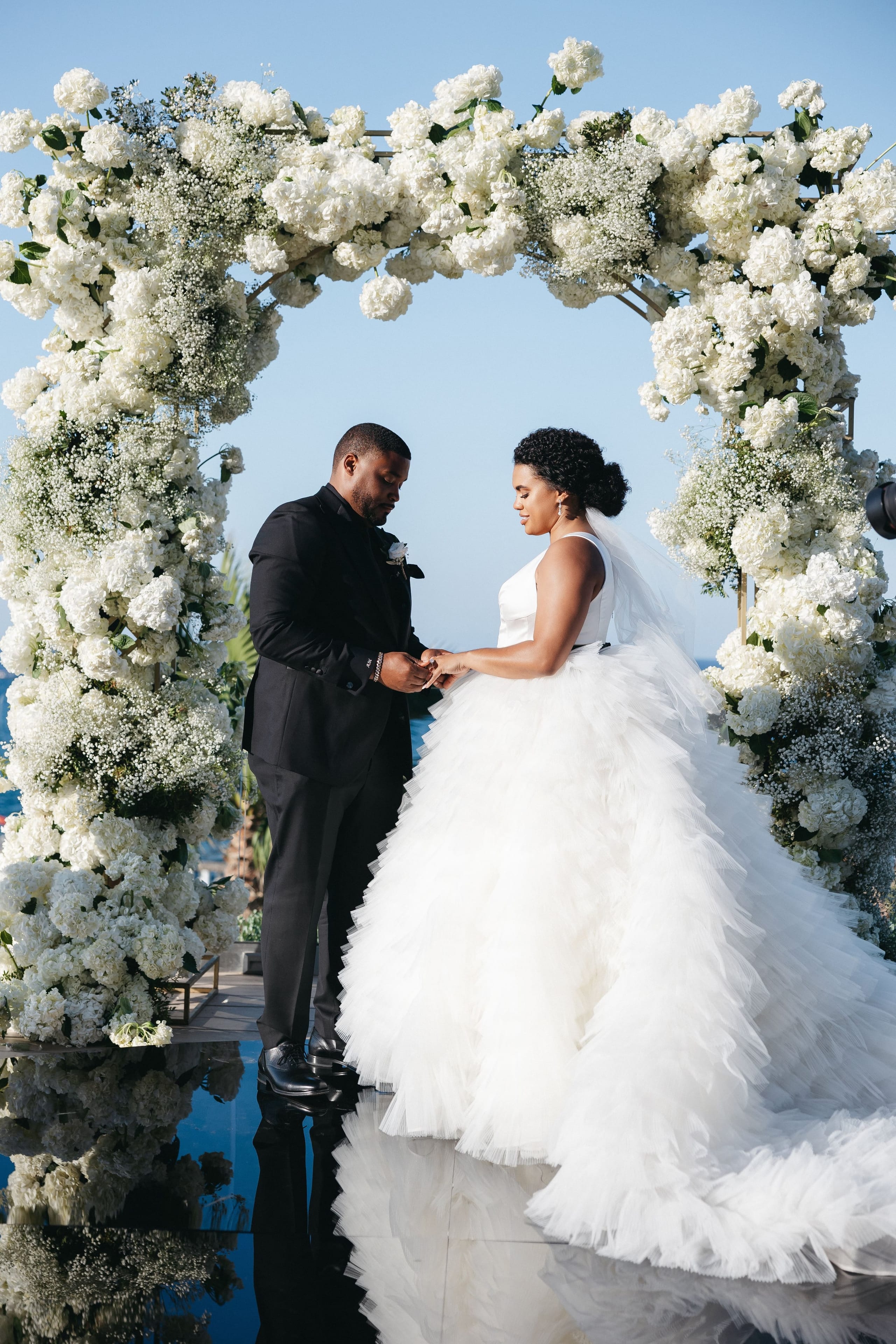 A bride and groom stand facing each other under a round arch completely covered in white blooms, mostly hydrangea and garden roses with airy sprays of baby's breath, at the Abaton Hotel in Greece. The groom, in a black suit with a white boutonniere, looks down as he slides a ring onto the bride's hand, while she gazes at him in her sleeveless gown with a tiered tulle skirt. Clear blue sky and a glimpse of the sea sit behind them, giving the scene a bright, romantic calm.