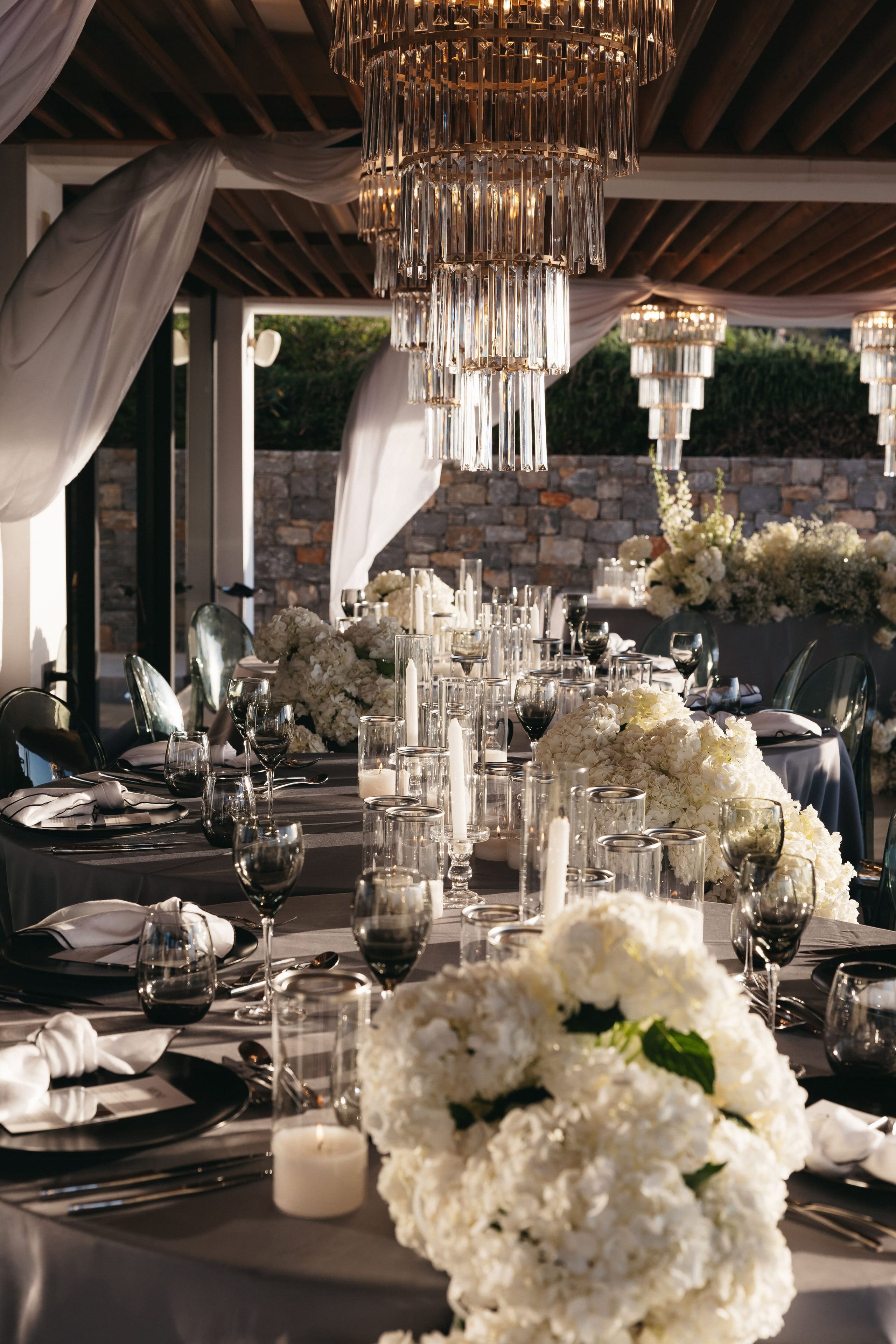 Long wedding banquet tables on a covered terrace at Abaton Island Resort & Spa in Crete, Greece, dressed in grey linens with smoky glassware and white napkins. Dense white hydrangea centrepieces sit along the tables, with tall pillar candles in glass hurricanes between them, and tiered crystal chandeliers hang from the timber ceiling above. Sheer white drapes frame the stone wall and greenery beyond, and the warm evening light gives the whole scene a soft, elegant glow.