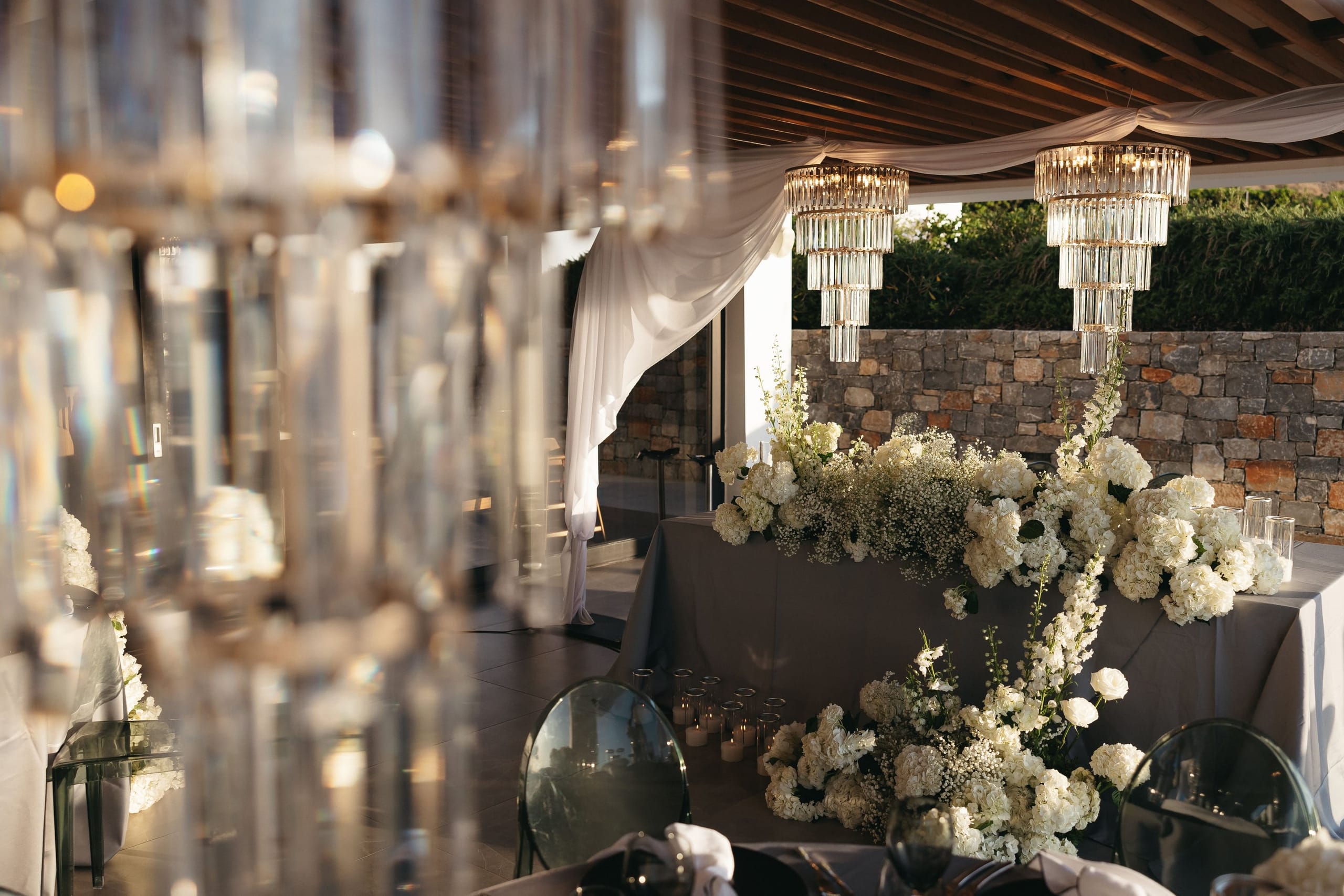 A wedding head table at Abaton Island Resort & Spa in Crete, Greece, dressed with a sweeping, low arrangement of white hydrangeas, gypsophila and tall white delphinium-like spires that spill down over a charcoal-grey tablecloth. Two tiered crystal chandeliers hang beneath a wooden pergola draped in soft white fabric, and glass chairs and candles glow in warm late-afternoon light against a rough stone wall and dark green hedging. Blurred crystal drops in the foreground add a soft, glittering frame to the scene.