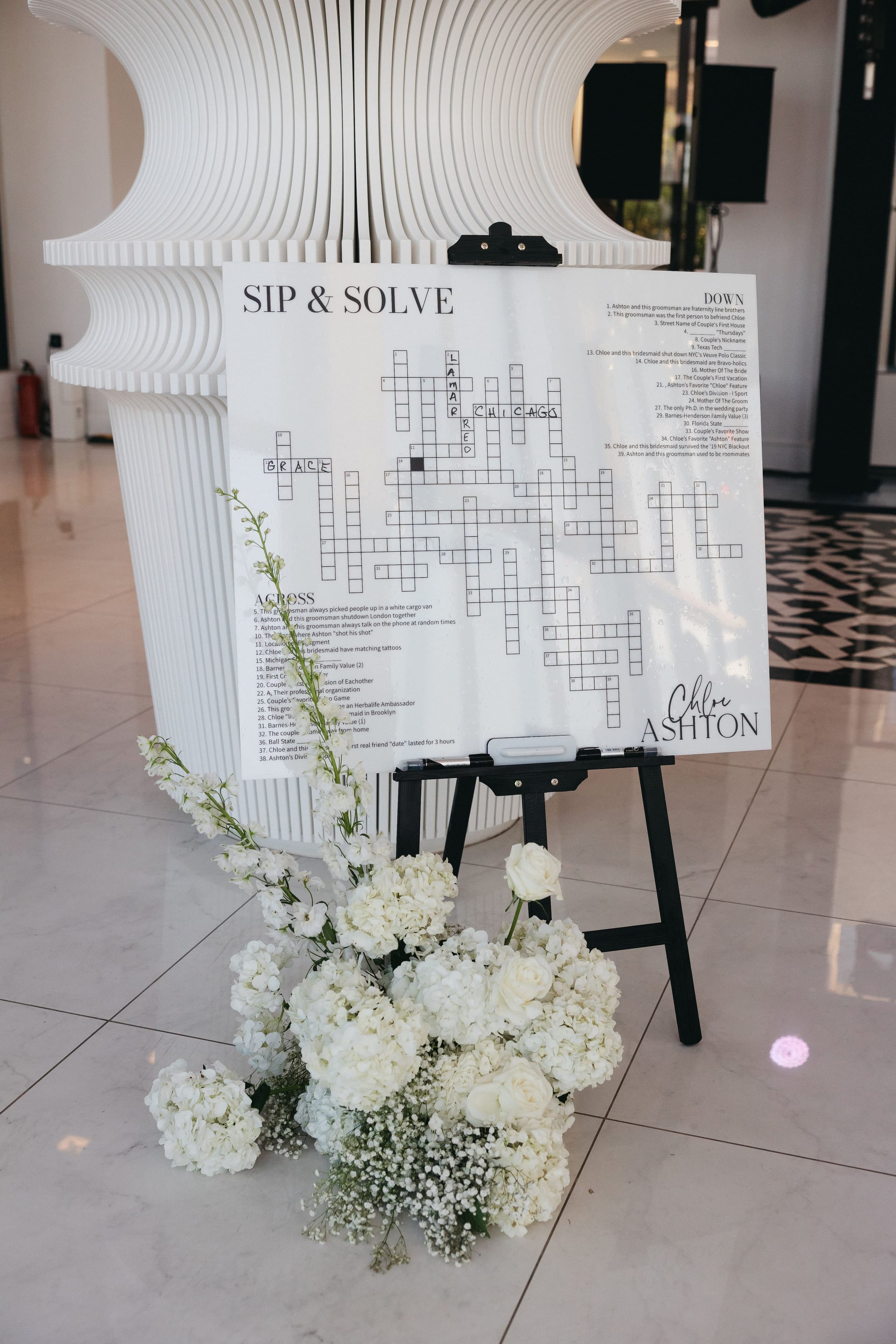 A white flower arrangement of hydrangeas, white garden roses and delicate baby's breath sits at the foot of a black easel holding a large crossword sign titled "Sip & Solve," set in the lobby of Abaton Island Resort & Spa on Crete, Greece, as part of a wedding celebration. The sign is black and white with clues in two columns, and the crossword grid features the words "Chicago," "Lamar" and "Brace" written in its squares. Behind it stands a sculptural white fluted column, and the glossy pale floor reflects the soft light.