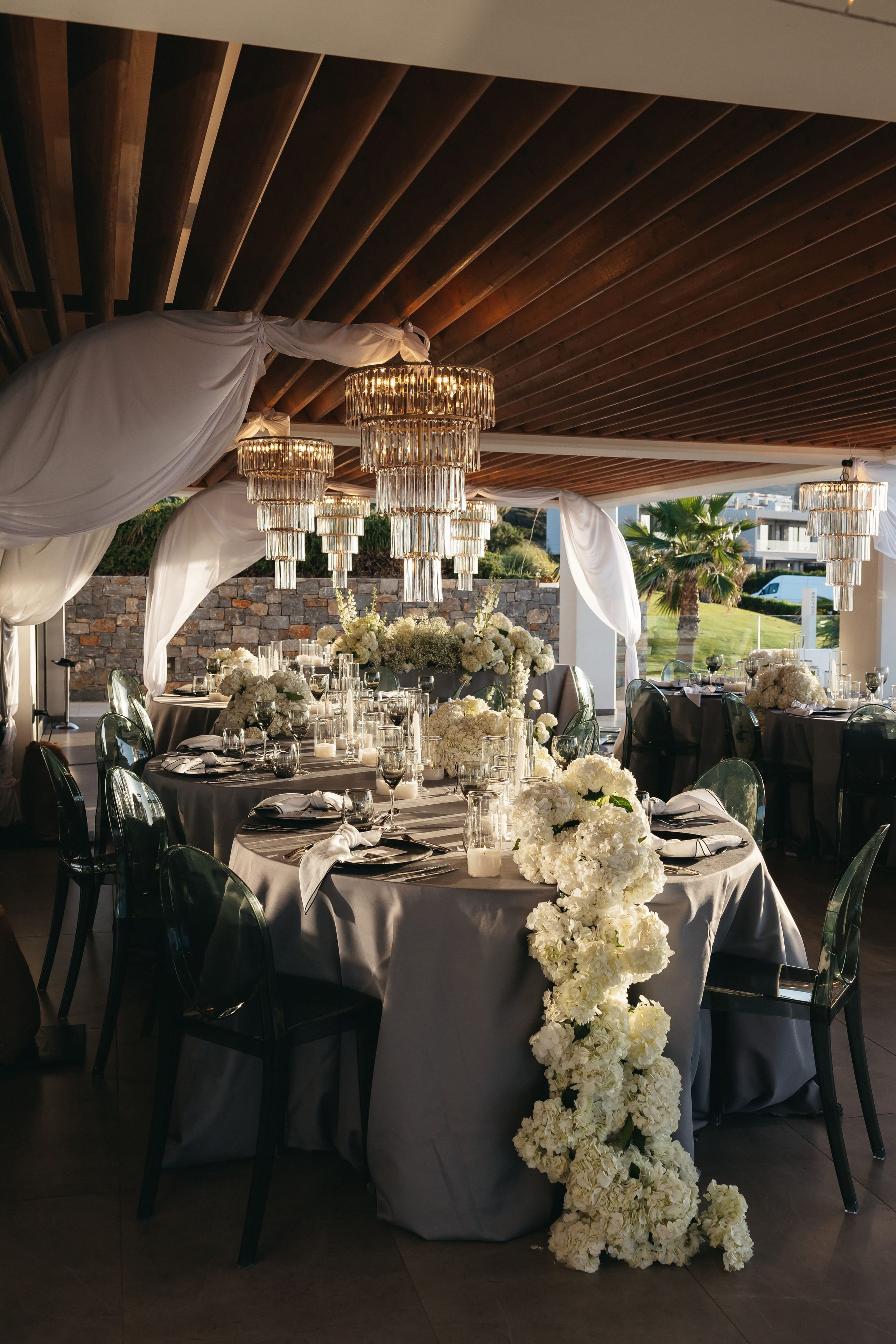 A wedding reception table at Abaton Island Resort & Spa in Crete, Greece, set with grey linen and dark glass chairs beneath tiered crystal chandeliers. A long garland of white hydrangeas trails down the side of the table, and white roses and gypsophila fill the low centrepieces, with pillar candles glowing among the glassware. Sheer white drapes frame the wooden-ceilinged terrace, and the warm evening light gives the scene a soft, elegant calm.