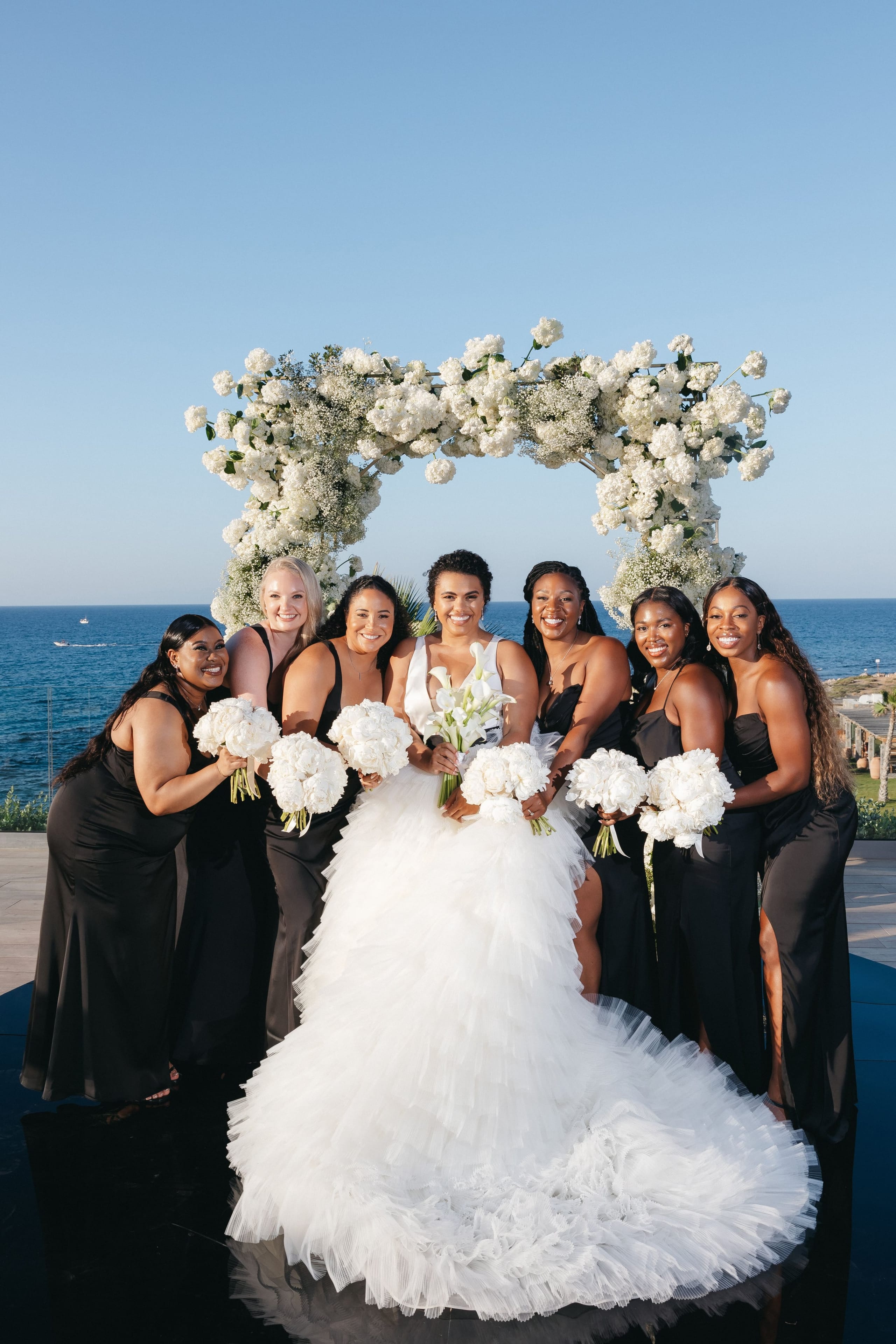 A bride in a voluminous white tiered tulle gown stands at the centre of six smiling bridesmaids in black satin dresses, each holding a white bouquet of peonies, roses and calla lilies, beneath a rectangular arch of white hydrangeas and baby's breath. Wedding flowers at Abaton Island Resort & Spa in Crete, Greece, with the deep blue Mediterranean sea and clear sky stretching behind them.
