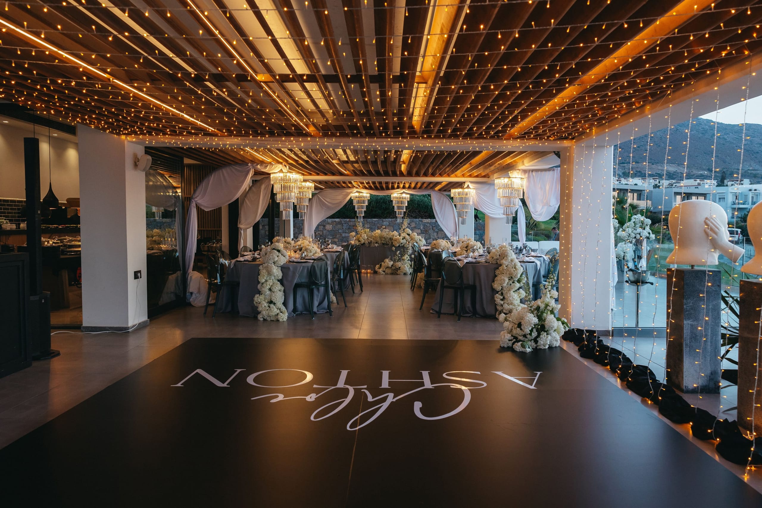 A wedding reception space at Abaton Island Resort & Spa in Crete, Greece, under a ceiling strung with warm fairy lights across wooden beams. Round tables draped in grey linen are dressed with tall white blooms, likely hydrangeas and garden roses, that trail down the tables and sit along the edges of the dance floor. A dark floor carries the mirrored names "Holly" and "Mark" in elegant script, and white drapes and crystal chandeliers add a soft, glowing finish.