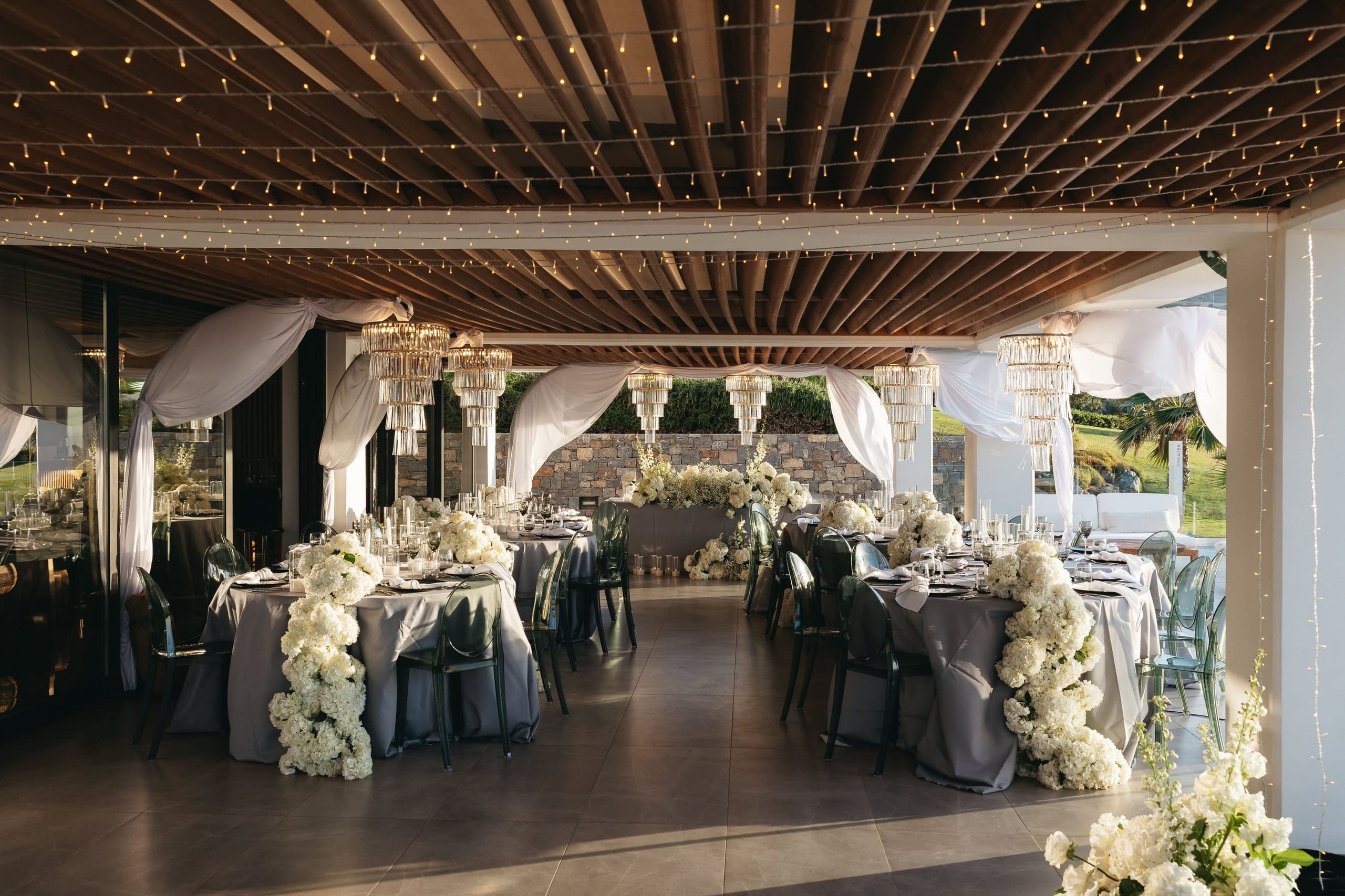 Long rectangular tables dressed in grey linen run through a pavilion strung with warm fairy lights beneath a slatted timber ceiling, with white drapes swagged between tiered crystal chandeliers. Cascades of white hydrangeas and white stock spill along the tables and gather in a sculptural arrangement at the far end, all set for a wedding at Abaton Island Resort & Spa on Crete, Greece.