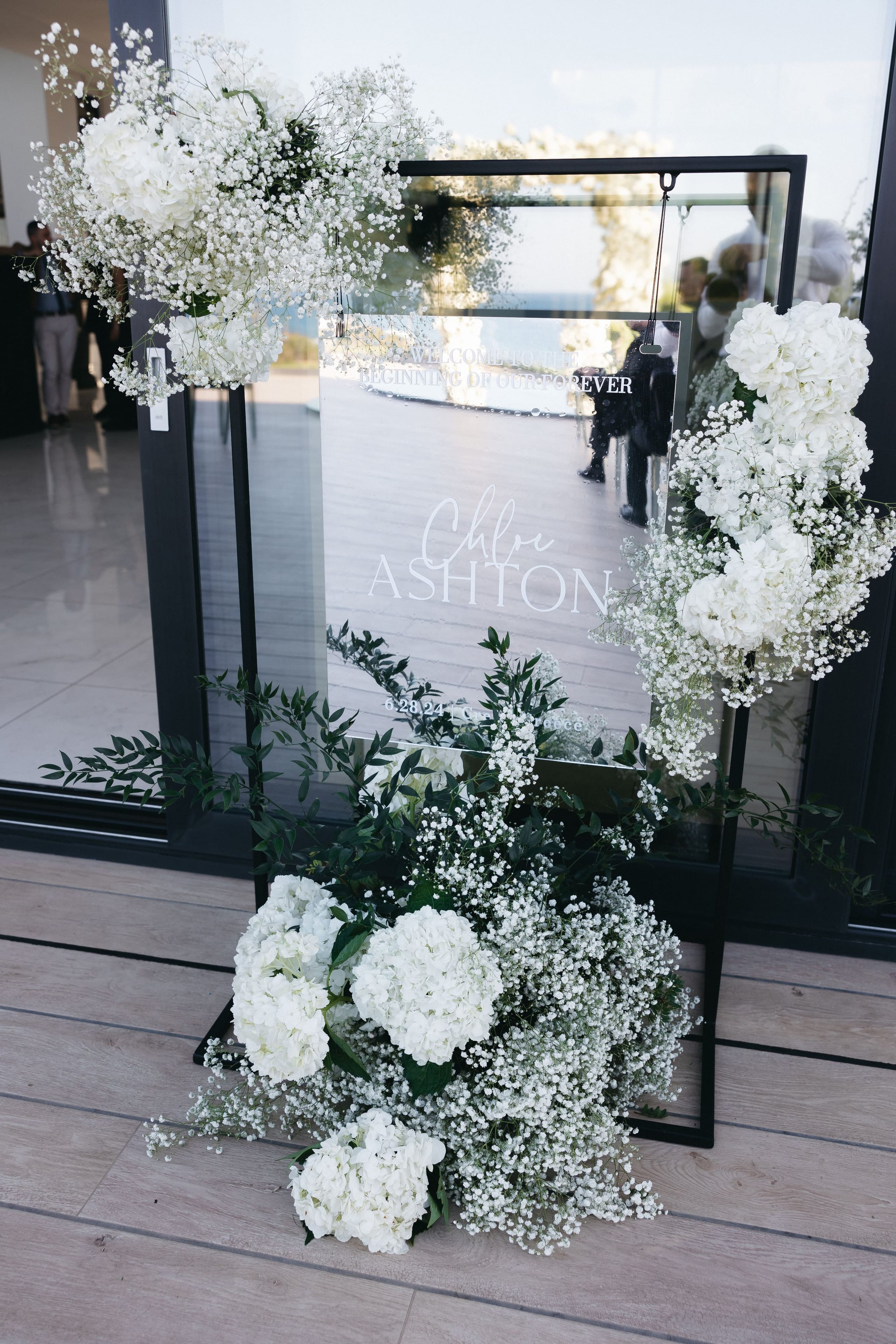 A black-framed mirror welcome sign at the entrance of a wedding in Crete, Greece, at Abaton Island Resort & Spa, dressed with all-white flowers: clusters of white hydrangeas and cascading baby's breath (gypsophila) tumble down both sides and gather at the base, with sprigs of green foliage weaving through. The mirror reflects the sea and guests, and the fine white lettering on its surface reads "Chloe Ashton" with a date below.