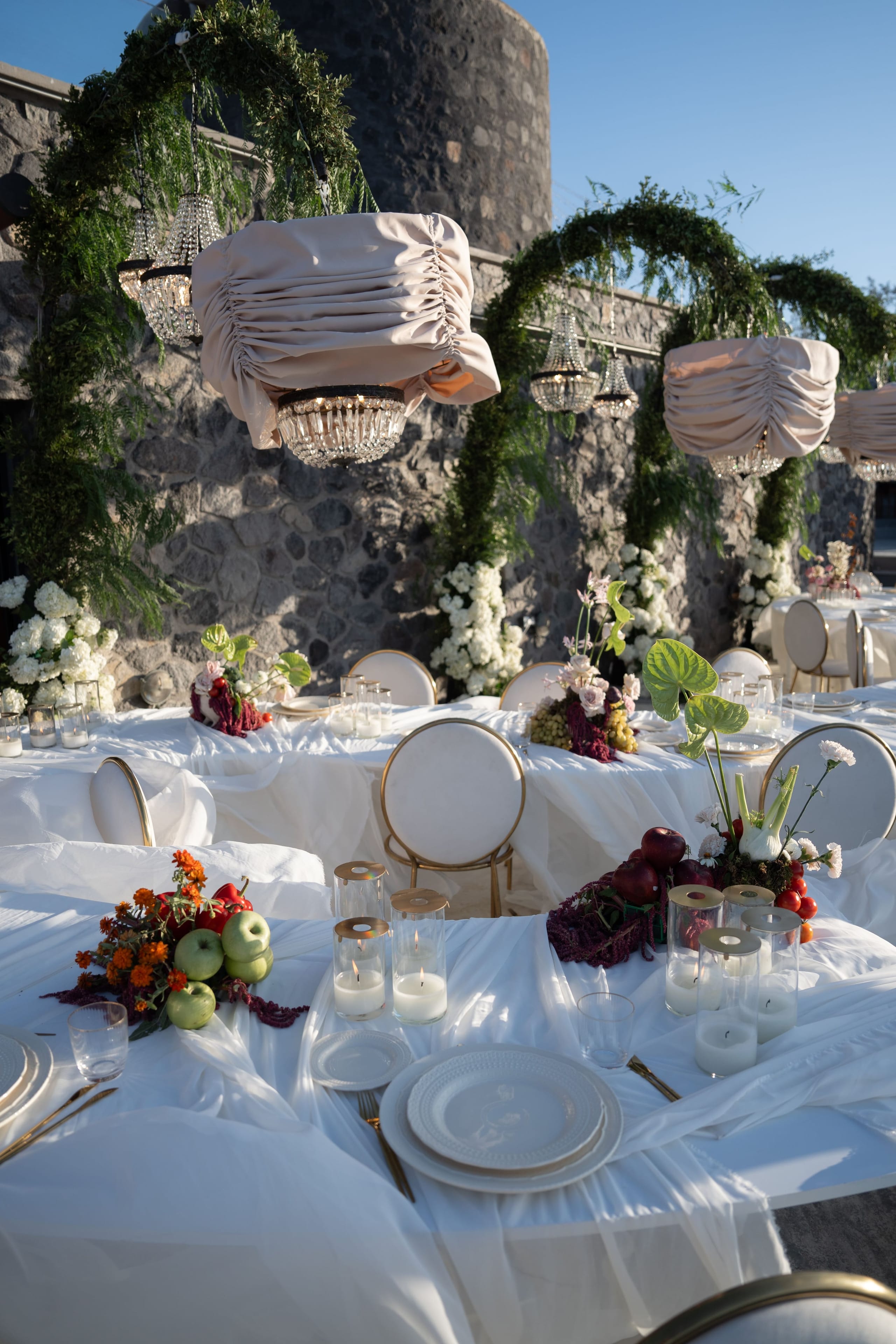 Long white tables dressed in sheer white linen and set with gold-rimmed plates sit beneath draped blush-pink chandeliers hung from curving garlands of trailing green foliage, with a rough stone wall behind. Low centrepieces of green apples, red apples, marigolds, fennel and deep red foliage, with white blooms, sit along the tables, lit by clusters of pillar candles in glass holders. This is a wedding table setting at Cavo Ventus Luxury Villa in Santorini, Greece.