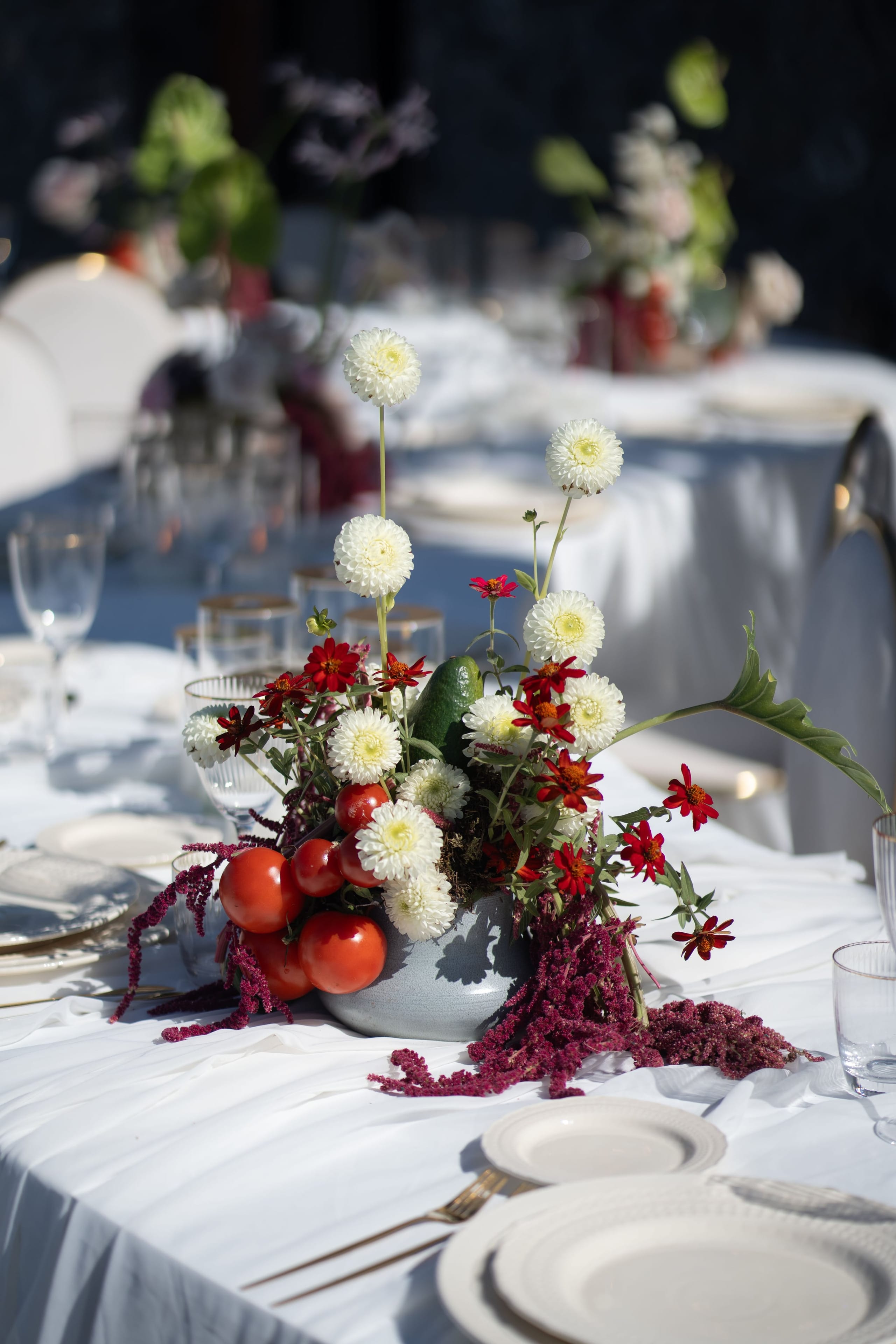 A low centrepiece on a white-linened wedding table at Cavo Ventus Luxury Villa in Santorini, Greece, arranged in a grey ceramic bowl. Round white pompom dahlias rise on long stems above small red zinnias, with trailing crimson amaranth spilling over the tablecloth, and bright red cherry tomatoes and a green pear-shaped fruit tucked among the blooms. Gold-rimmed glasses and white plates sit softly blurred behind it in a long, sunlit row.