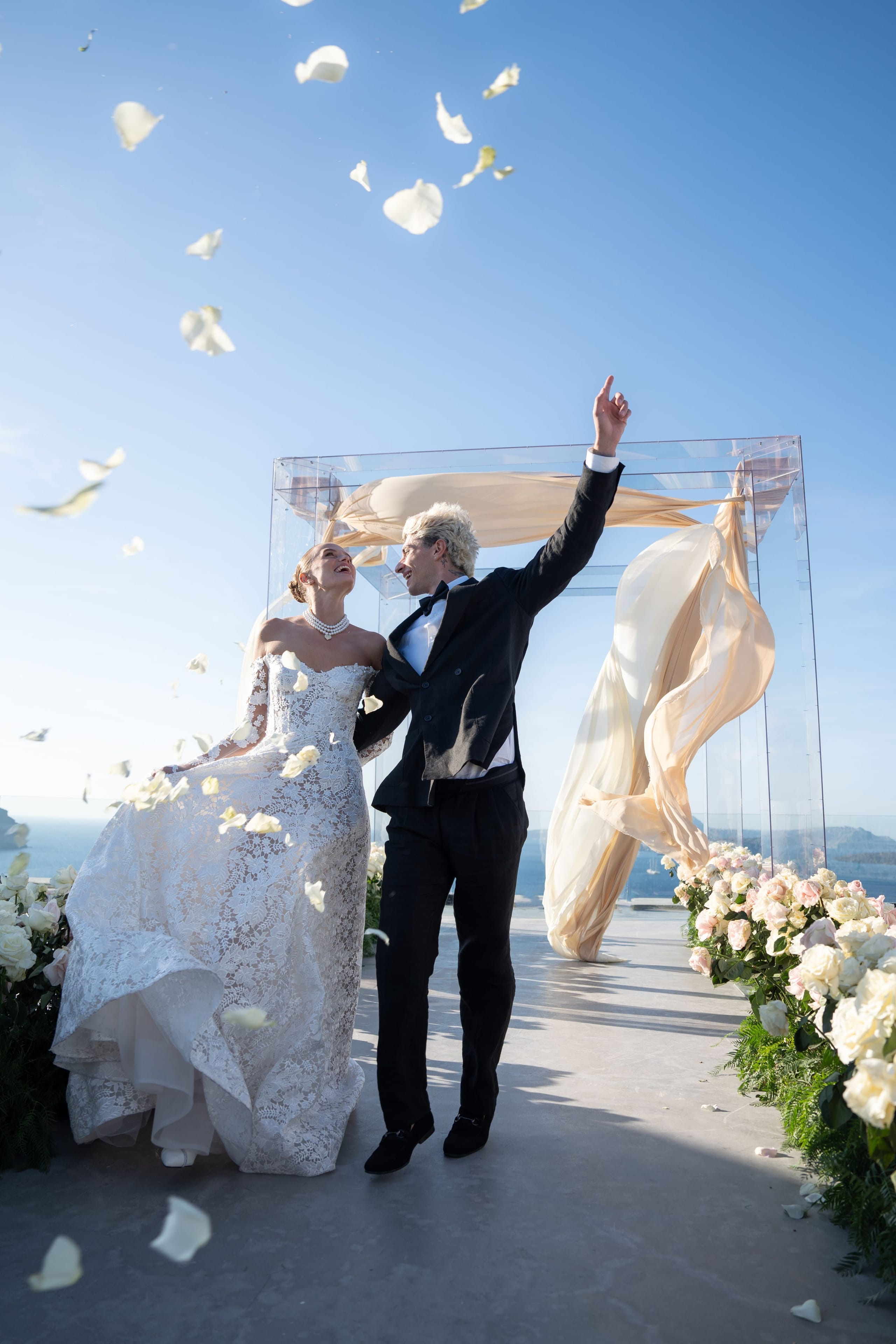 A bride in a white lace gown with a pearl necklace and a groom in a black suit with a bow tie walk together along a terrace, laughing as white rose petals swirl through the bright blue sky. Behind them stands a clear acrylic arch draped with flowing champagne-coloured fabric, and pale garden roses and white blooms line the path, all for a wedding at Cavo Ventus Luxury Villa in Santorini, Greece.
