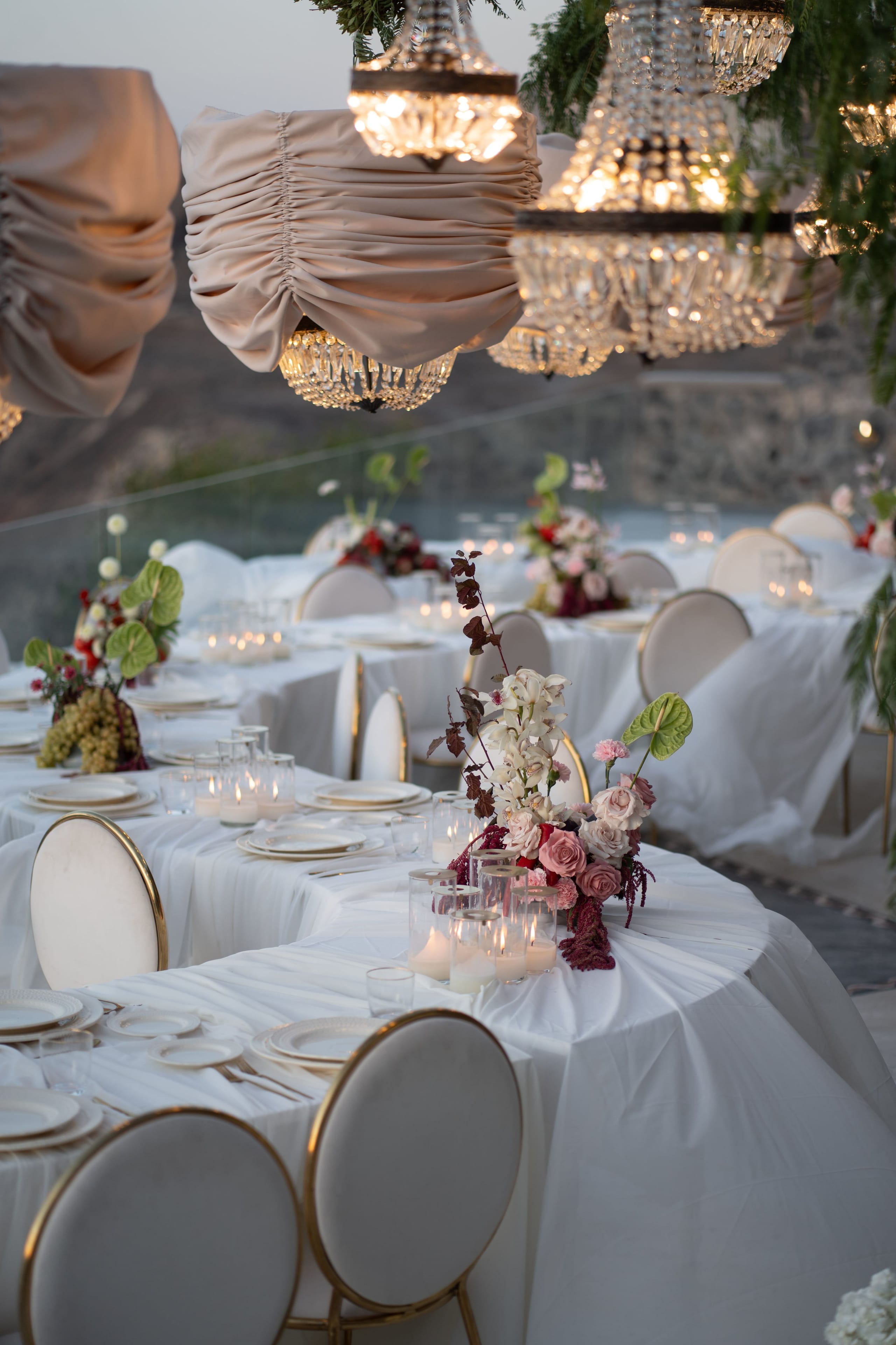 Long banquet tables draped in white linen stretch toward a glass balustrade at dusk, lit by crystal chandeliers hanging beneath blush fabric shades. Each place is set with white plates and gold-rimmed round chairs, and low centrepieces of blush roses, white orchids, green anthurium and deep burgundy foliage sit among flickering pillar candles. This is a wedding table setting at Cavo Ventus Luxury Villa in Santorini, Greece, with the calm evening sea beyond.