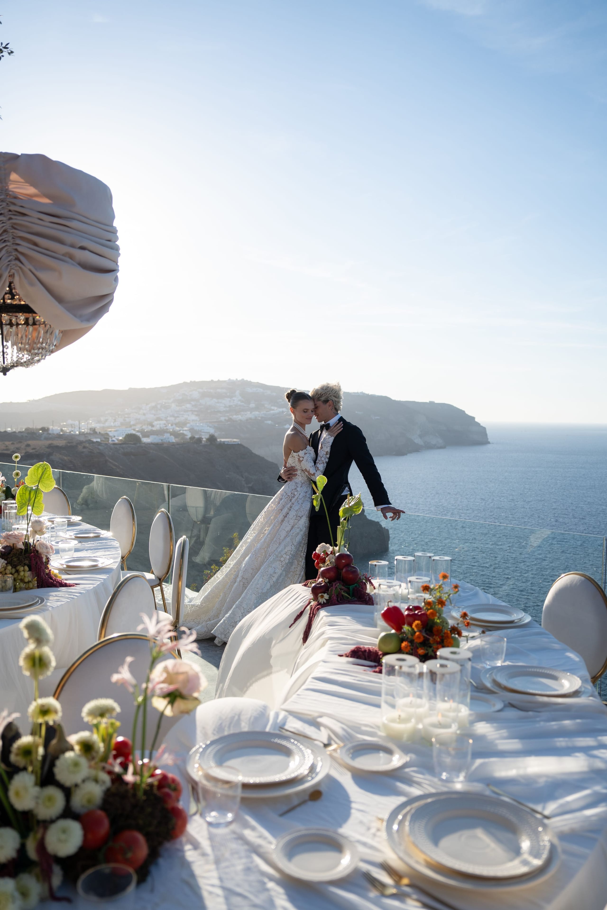 A bride in a lace gown and a groom in a black tuxedo stand close together at the edge of a clifftop terrace at Cavo Ventus Luxury Villa in Santorini, Greece, with the blue Aegean sea and a white-washed village stretching behind them. The long wedding table in front of them is dressed in white linen with gold-rimmed plates, glowing pillar candles, and low centrepieces of white ranunculus, pink blooms, red tomatoes and green anthurium leaves. Warm late-afternoon sunlight softens the scene into a calm, romantic mood.