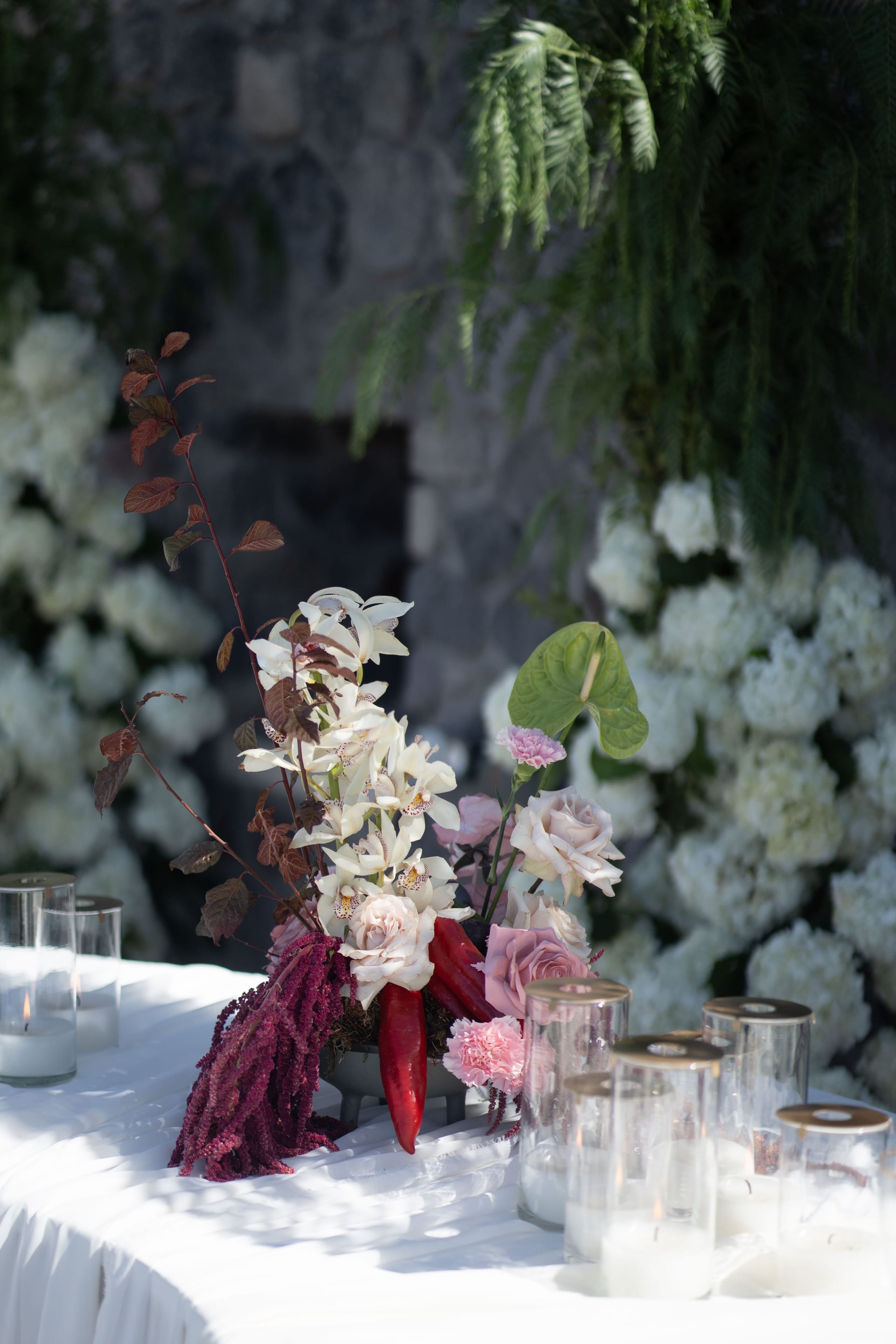 A low, asymmetrical wedding table centrepiece on a white draped table at Cavo Ventus Luxury Villa in Santorini, Greece, built around white cymbidium orchids, blush garden roses and a pale pink carnation. Trailing burgundy amaranthus spills down one side, with deep red chili peppers, a green anthurium and slender bronze-brown foliage rising behind. Glass candle hurricanes with gold lids sit beside it, and soft shadows fall across the cloth in the daylight, while a wall of white hydrangeas and dark stone glows behind.