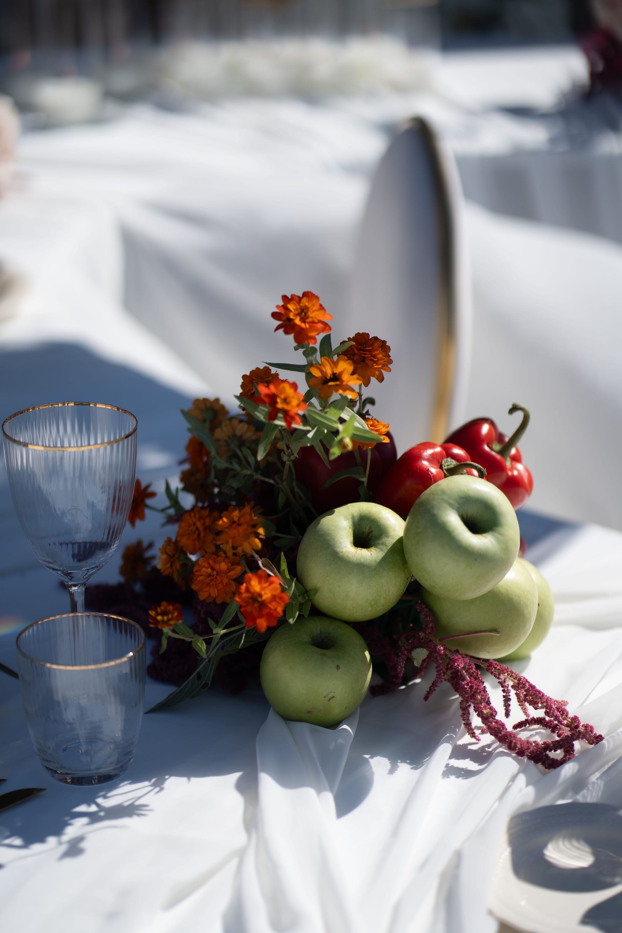 A wedding table centrepiece on a white linen cloth in Santorini, Greece, at Cavo Ventus Luxury Villa. Small orange marigold-style blooms and sprigs of green foliage rise from a cluster of green apples, with a red bell pepper tucked behind and trailing magenta amaranth spilling across the cloth. Beside it sit two clear ribbed glasses with thin gold rims, and the bright sunlight casts soft shadows across the table.