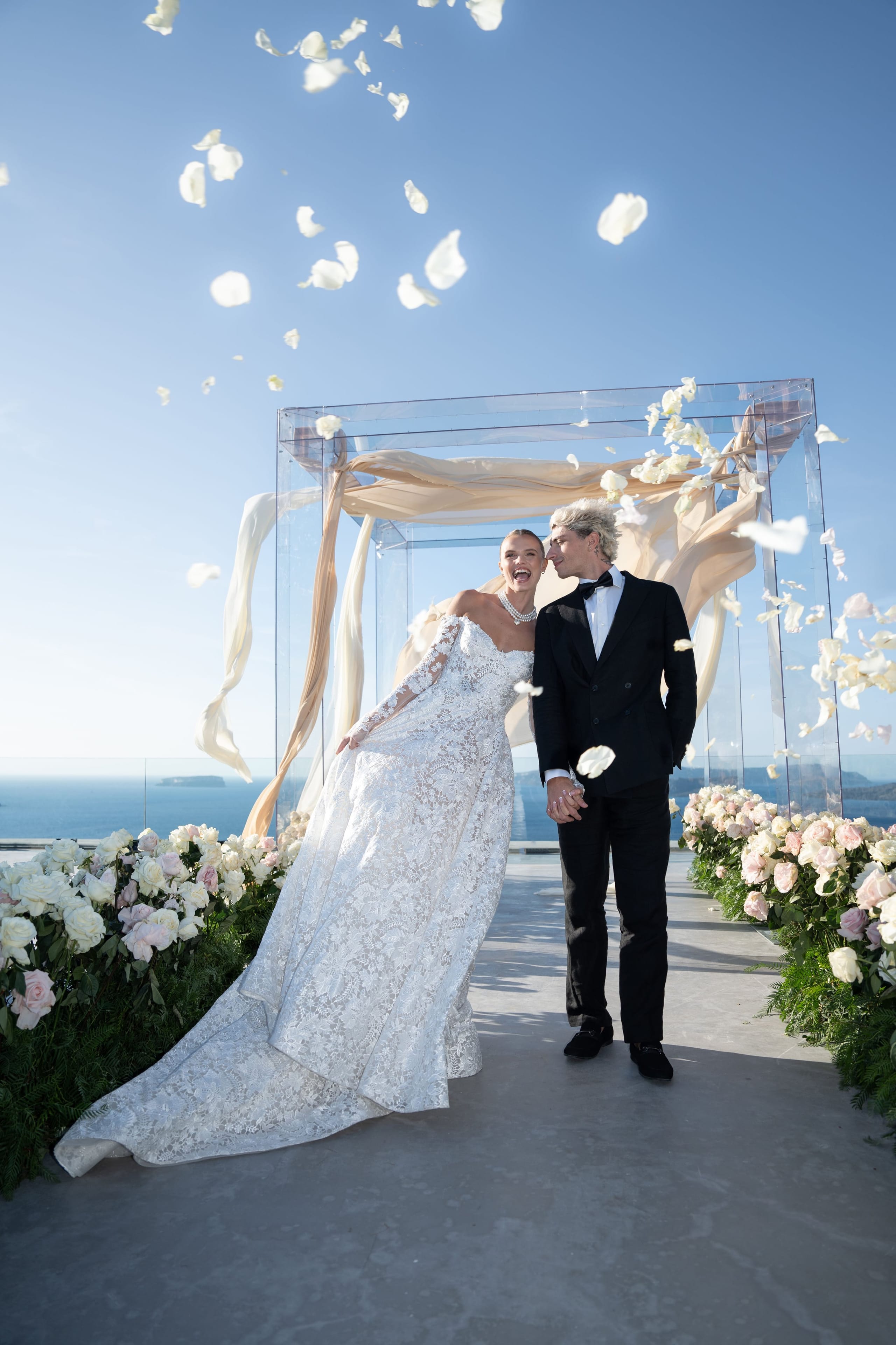 A bride in a lace gown with a long train laughs brightly as she holds up her skirt, walking beside a groom in a black tuxedo and bow tie along an aisle lined with white and blush roses and fern. Petals drift through the air beneath a draped cream fabric canopy, with the Aegean Sea glittering behind them at Cavo Ventus, a wedding venue in Greece, where these are wedding flowers for an outdoor ceremony.