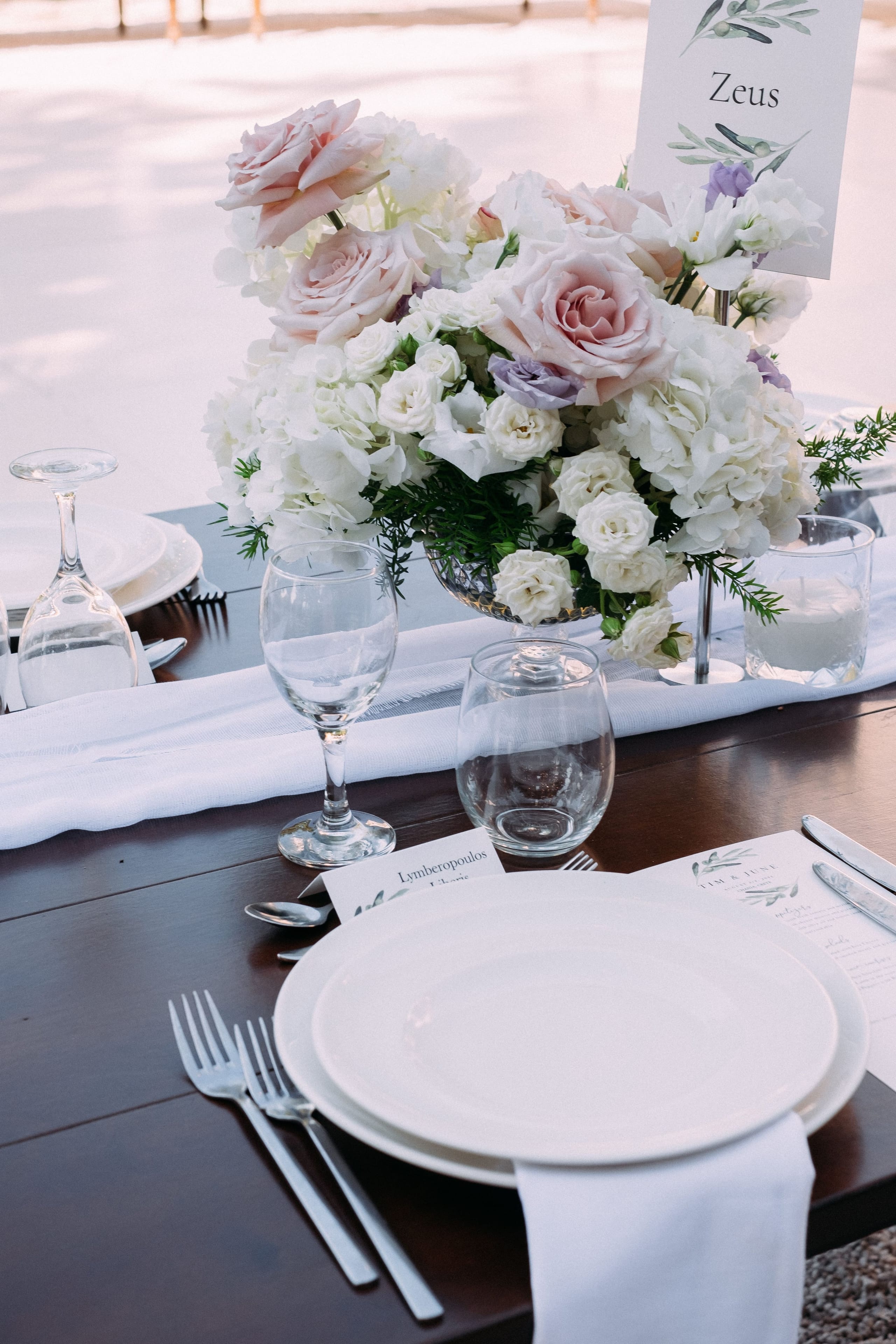 A wedding table setting at Dourakis Winery in Alikampos, Crete, Greece, with a low centrepiece of blush-pink garden roses, white hydrangeas, white spray roses and small lavender-purple blooms, softened with feathery green sprigs. The white plates, clear stemware and polished cutlery sit on a dark wooden table, with a white linen runner beneath the arrangement and a menu card and name card placed beside the plates.