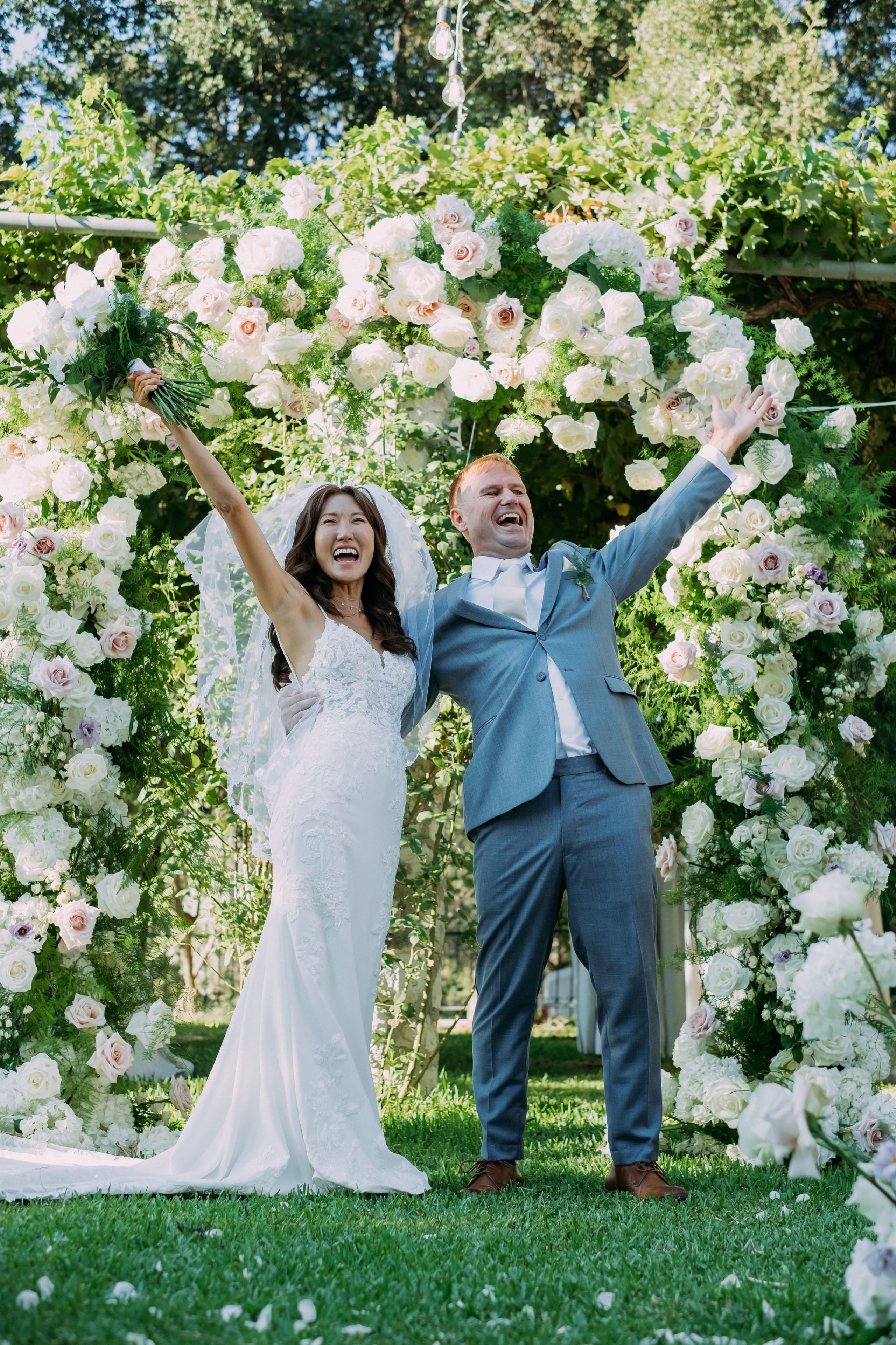 Two newlyweds laugh joyfully with their arms raised in front of a lush wedding arch of white garden roses, blush roses and hydrangeas, softened by feathery green fern fronds and climbing vines at Dourakis Winery in Greece. The bride, in a fitted lace gown and sheer veil, holds a green bouquet high in one hand, while the groom wears a dusty blue suit with a light tie and a small sprig on his lapel. Dappled sunlight falls across the flowers, creating a bright, celebratory mood.