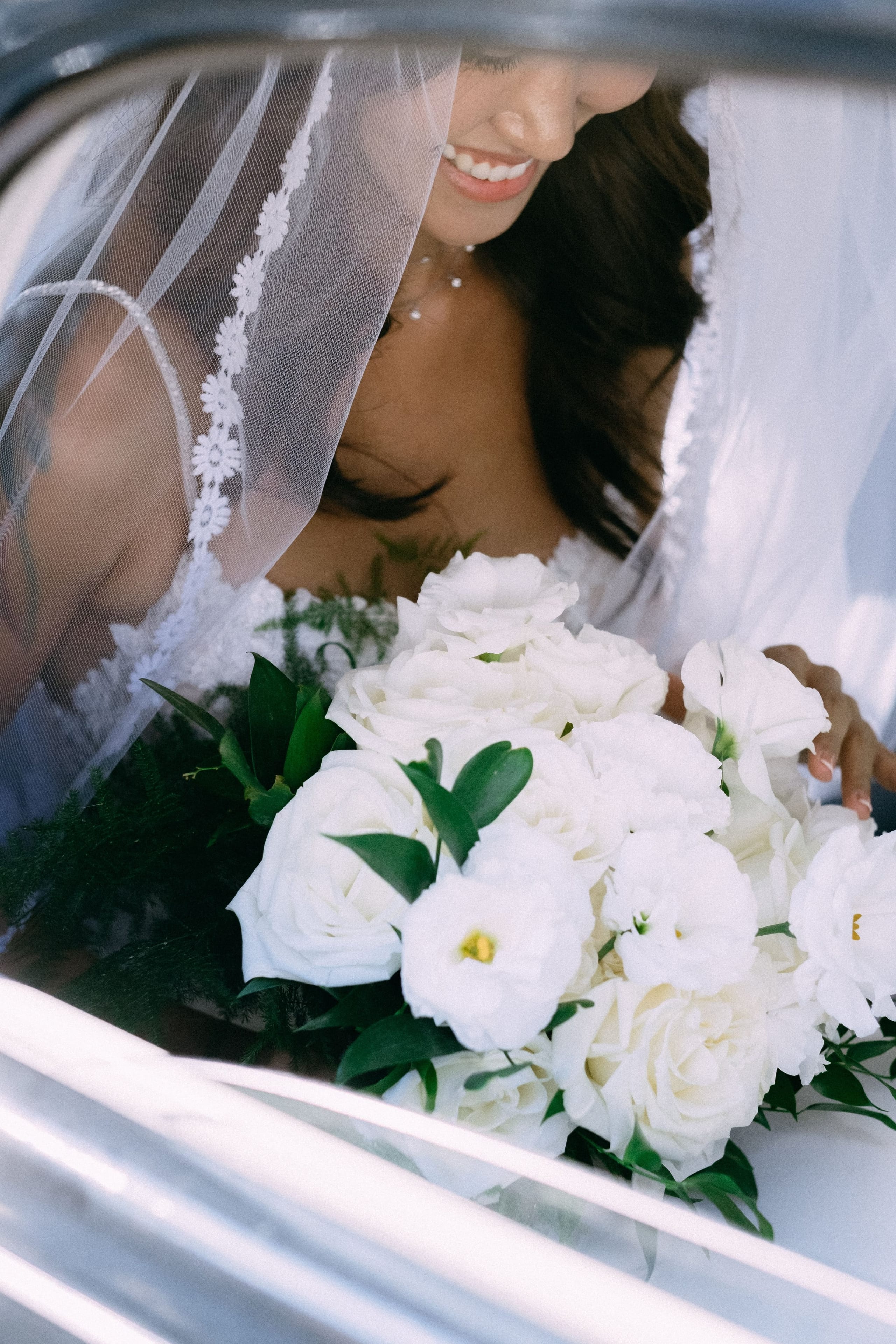 A bride smiles down at her bouquet through a sheer lace-edged veil while seated in a white car, her hand resting on the window frame. The bouquet is a full, rounded arrangement of white lisianthus and garden roses with glossy dark green leaves and sprigs of feathery fern, a wedding flower design for her big day at Dourakis Winery in Alikampos, Crete, Greece.