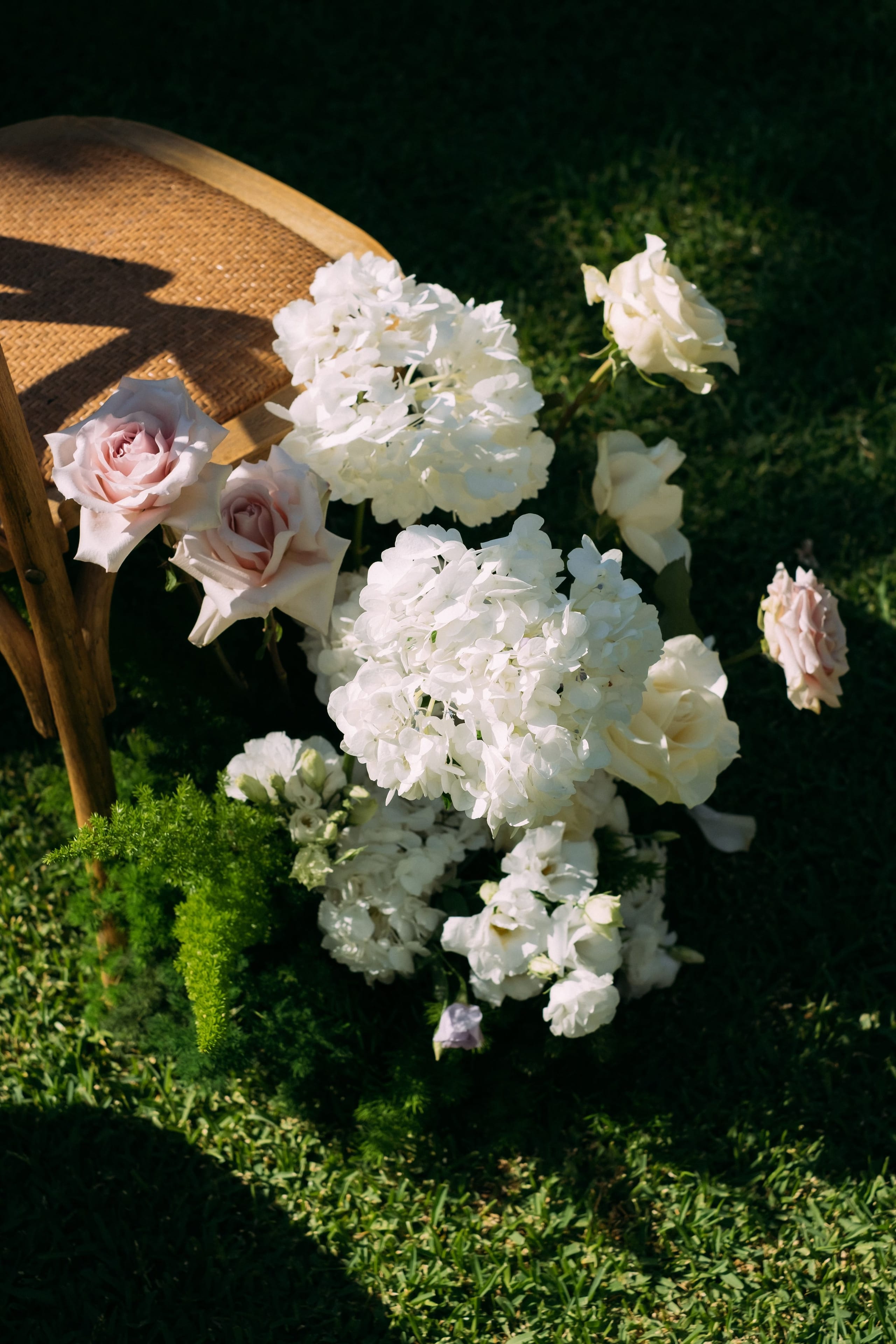 A low cluster of white hydrangeas, cream garden roses and blush-pink roses spills across the grass at the foot of a woven rattan chair, part of the wedding flowers at Dourakis Winery in Alikampos, Crete, Greece. Sunlight falls across the scene, casting the chair's shadow on the lawn, and soft green fern-like foliage tucks in beneath the blooms. The arrangement feels airy, romantic and relaxed, as if it had been left casually among the garden grass.