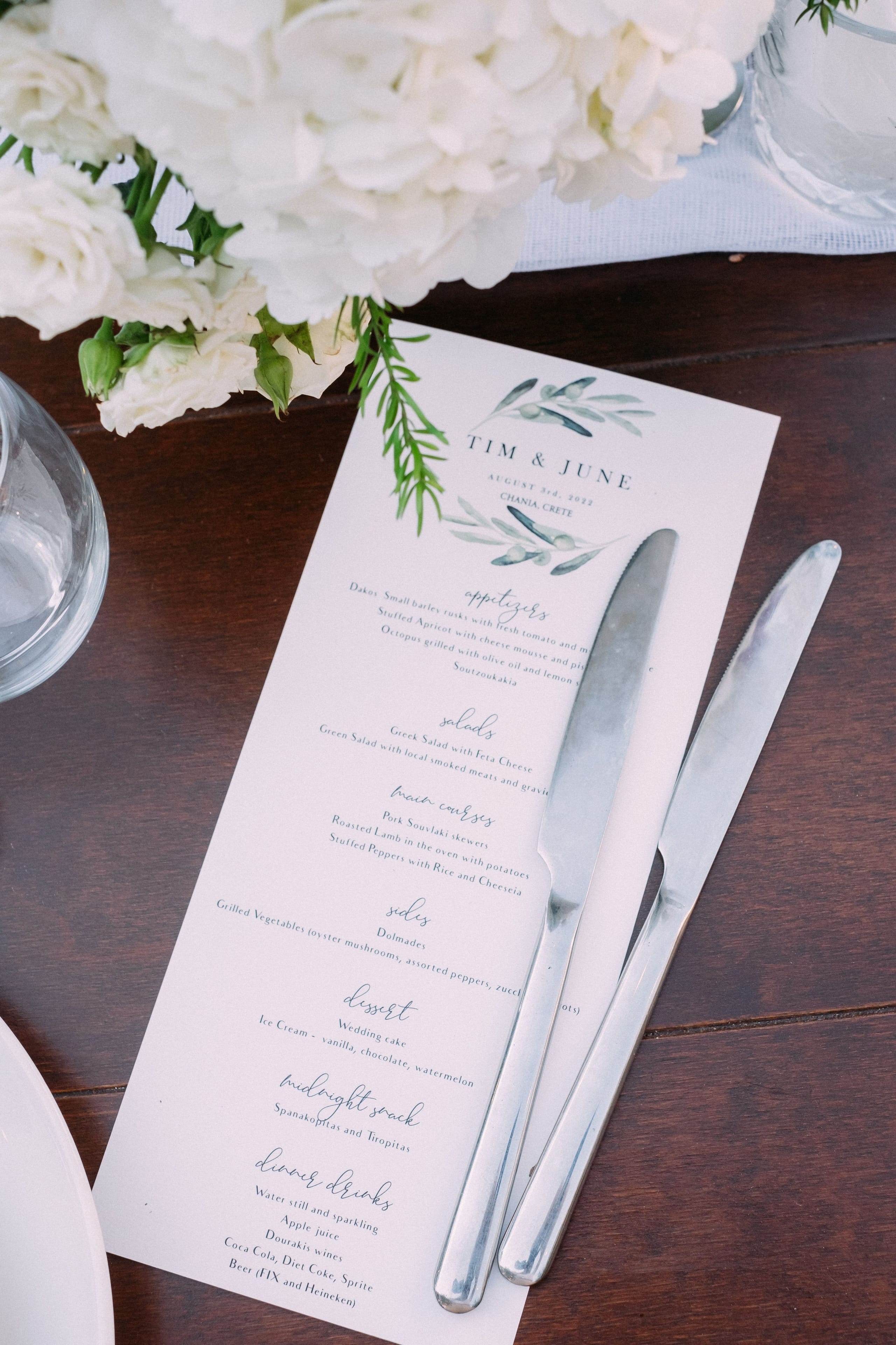 White hydrangeas and white lisianthus with green buds spill across the top of a wedding table at Dourakis Winery in Alikampos, Crete, Greece. Beneath them lies a cream menu card with a painted olive-branch wreath, reading Tim & June, and two polished silver knives rest across its text on a dark wooden table. The soft, natural light and muted palette give the scene a calm, elegant feel.