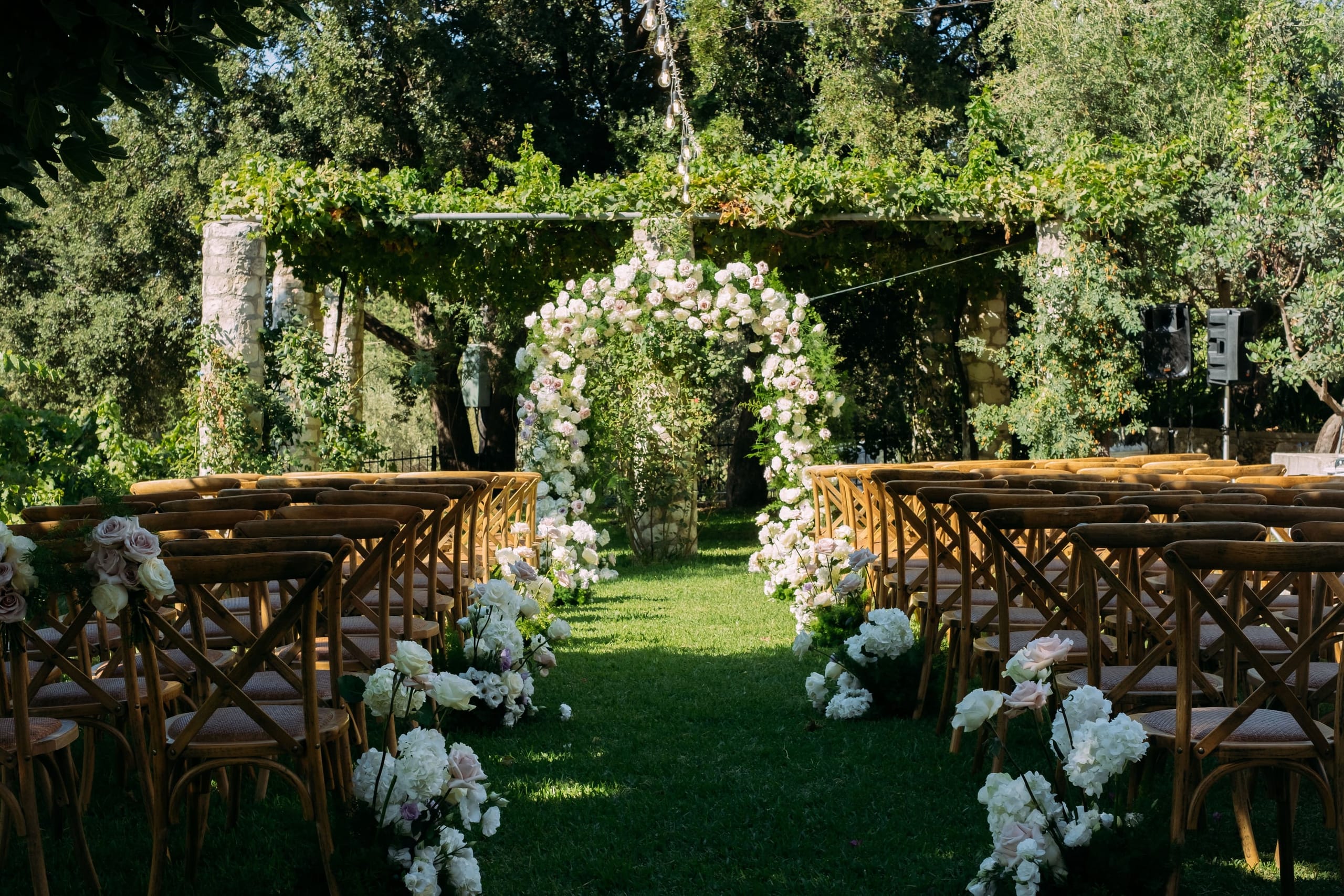 A wedding ceremony aisle on a grassy lawn at Dourakis Winery in Alikampos, Crete, Greece, lined with rows of wooden cross-back chairs. A full ceremony arch of white and blush roses and other soft white blooms stands at the far end, framed by a leafy vine-covered pergola and ancient stone columns. Clusters of white hydrangeas and pink-tinged roses decorate the aisle, and strings of lights hang overhead beneath the dappled shade of the trees.