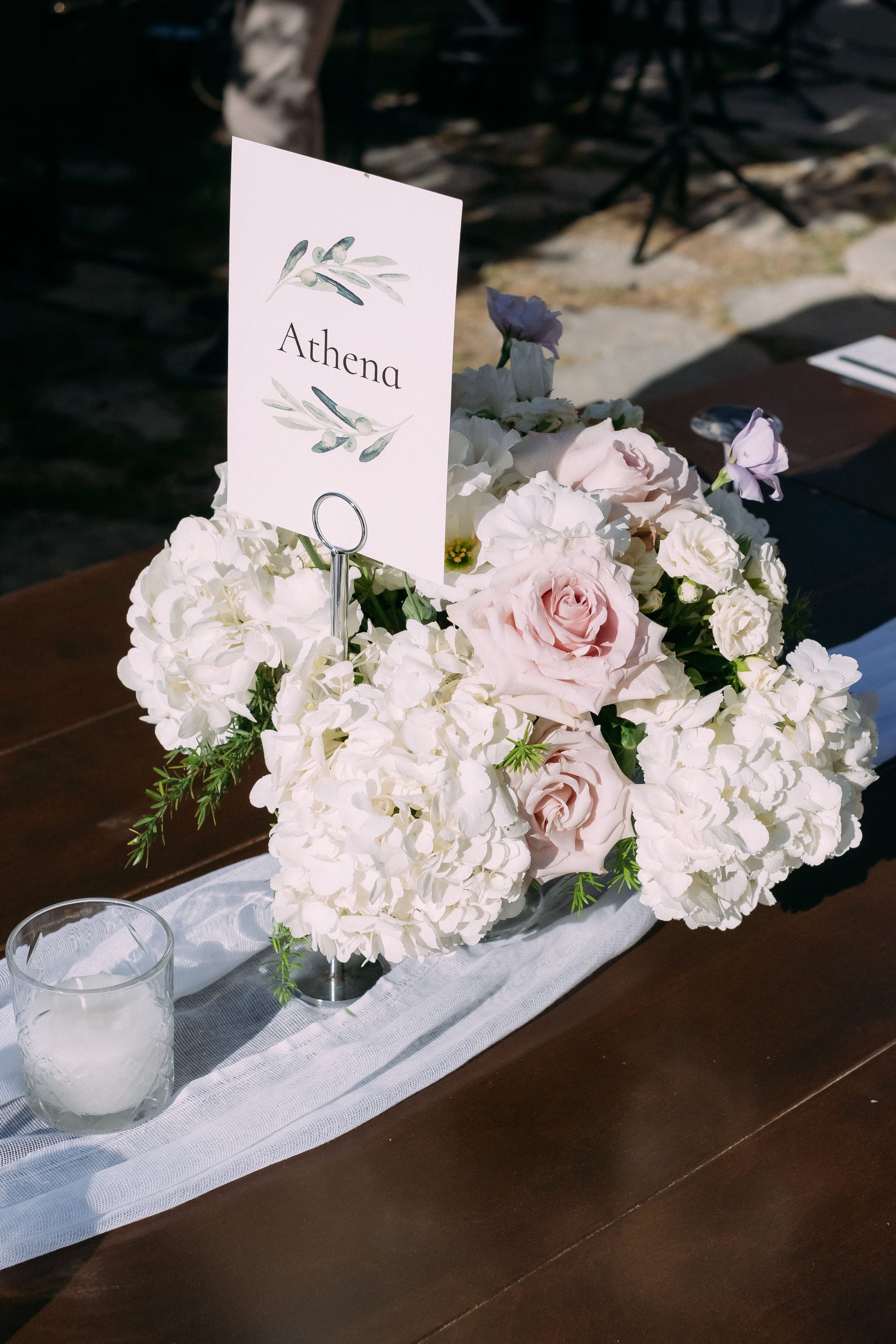 A low centrepiece of white hydrangeas, blush garden roses, small white spray roses and sprigs of green rosemary sits on a dark wooden table at a wedding at Dourakis Winery in Alikampos, Crete, Greece. A slim white place card with olive branch sprigs and the name Athena is propped in a silver holder above the blooms, and a sheer white table runner and a frosted glass votive lie beside it. Soft natural light falls across the scene.