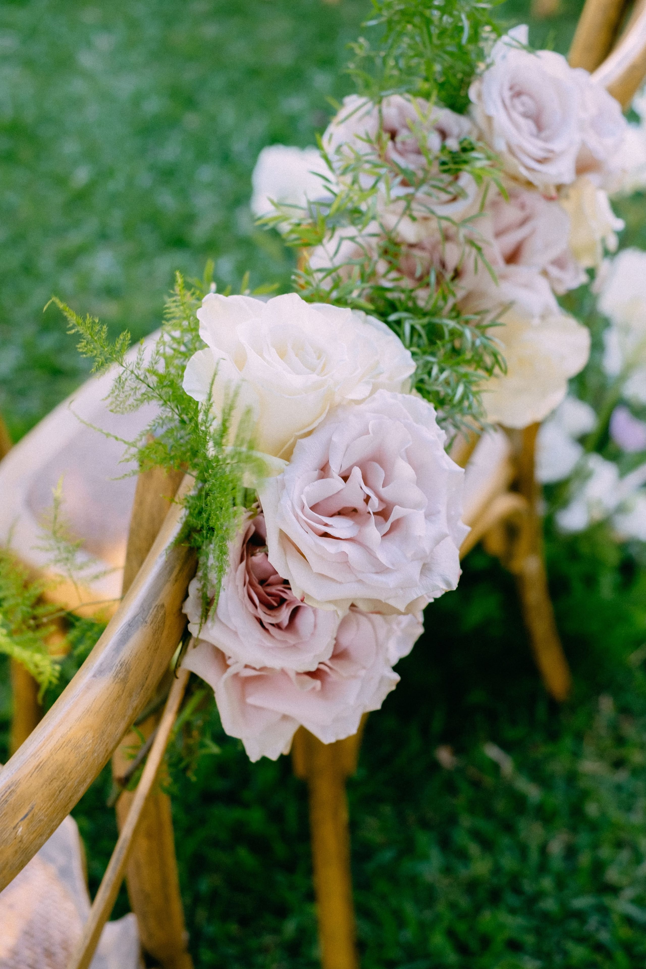 Pale blush and cream garden roses, tucked with feathery green asparagus fern, are tied to the wooden crossback chairs of a wedding aisle at Dourakis Winery in Alikampos, Crete, Greece. The soft pink and ivory blooms stand out against the lush green lawn, which curves away softly out of focus behind them. The gentle, natural light gives the scene a calm, romantic feel.