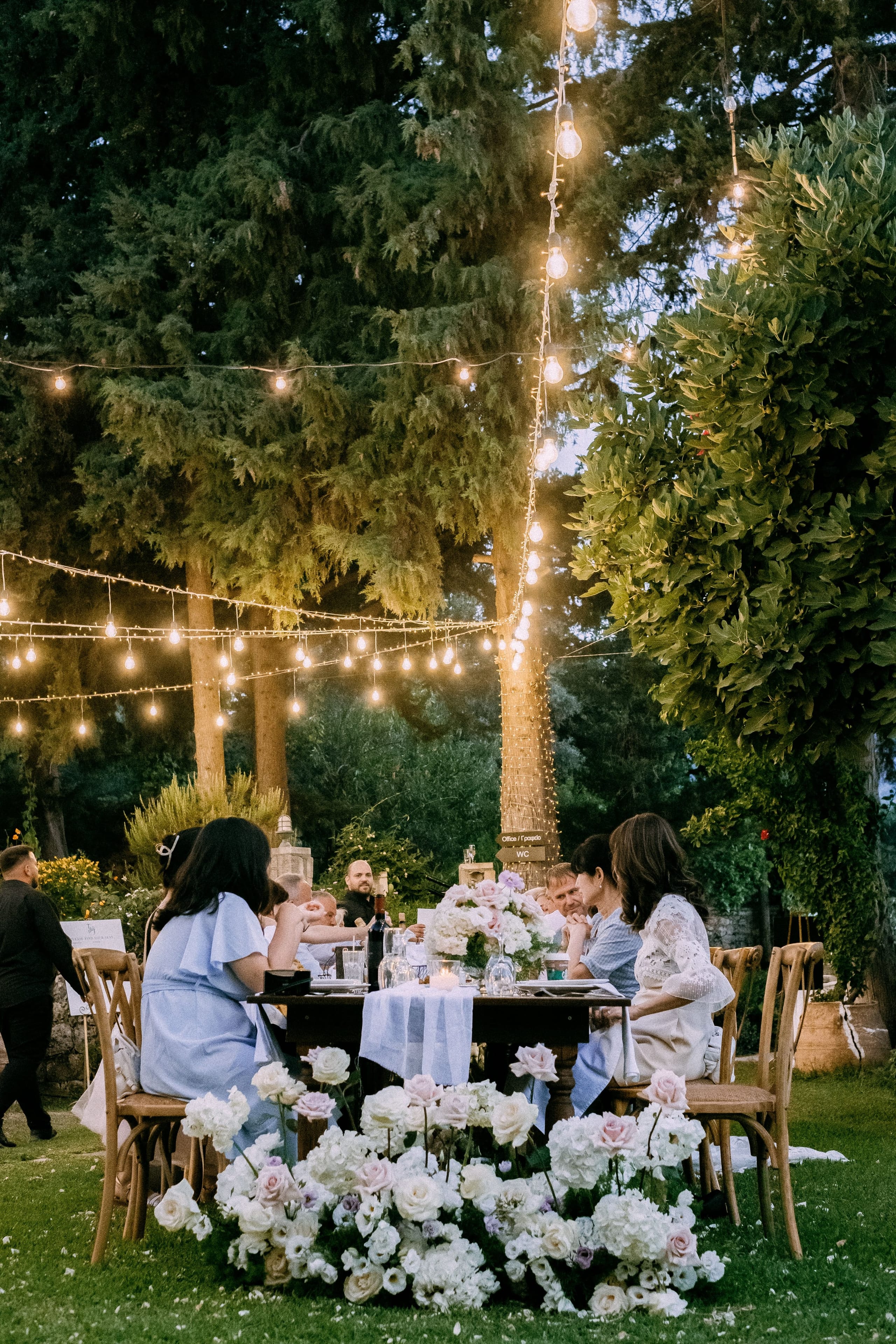 Low white and blush floral arrangements in pale pink, cream and lavender sit along the grass at the foot of a long wooden table, where guests in soft blue and white dresses dine under a canopy of warm string lights strung through tall cypress trees. This wedding table setting for June and Tim is at Dourakis Winery in Alikampos, Crete, Greece, with the bulbs glowing against the dusk-blue sky.