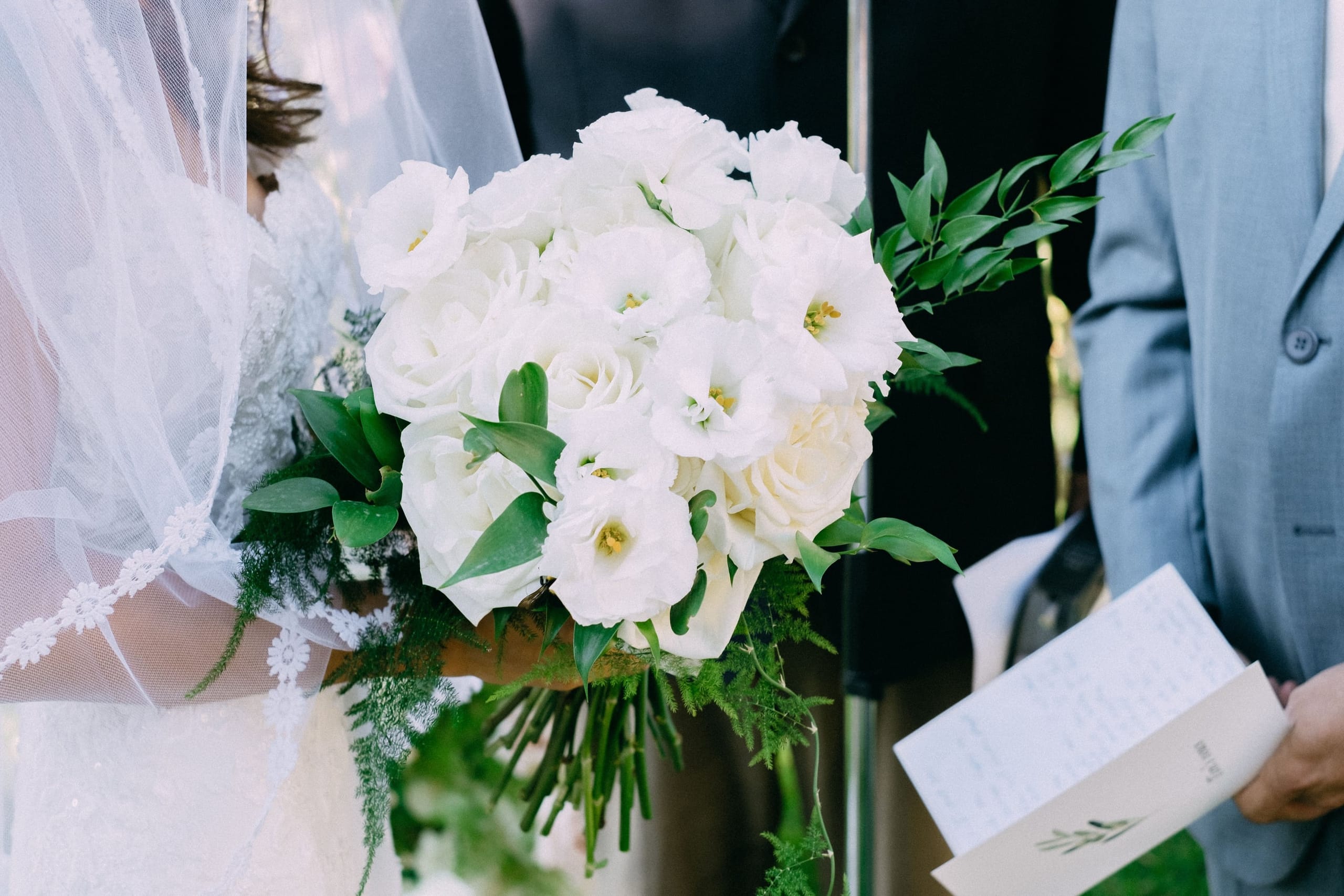 A bridal bouquet of white lisianthus and creamy garden roses, with sprigs of green leaves and feathery asparagus fern, held by a bride in a lace-edged veil at Dourakis Winery in Alikampos, Crete, Greece. Beside her, a man in a light grey suit holds a small printed ceremony programme, and the soft, natural light gives the whole scene a calm, airy feel.