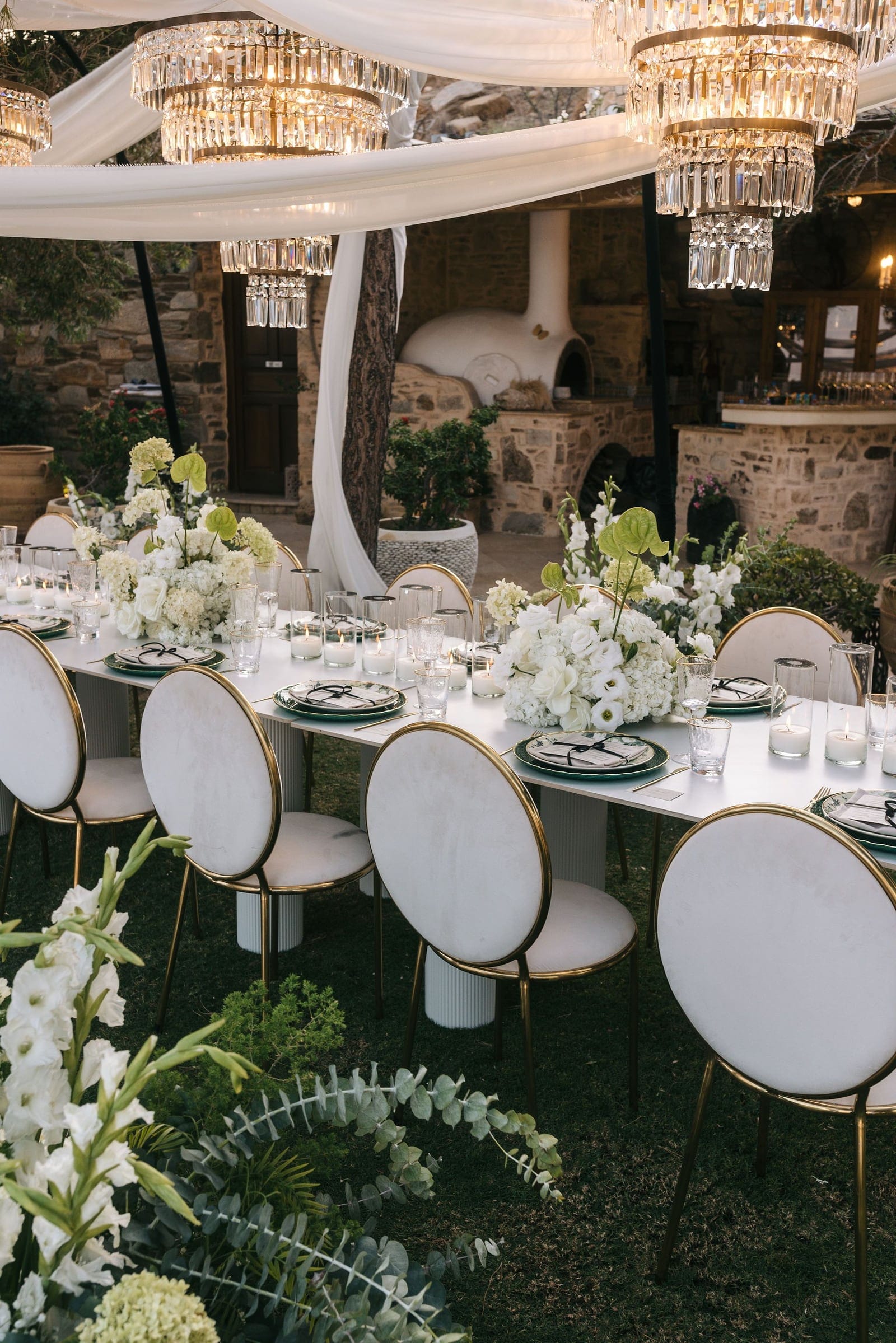 A long white dining table under a canopy of billowing white fabric and tiered crystal chandeliers, set for a wedding dinner in the garden of Cycladic Gem Luxury Villa on Ios Island, Greece. Low centrepieces of white hydrangeas, roses and lime-green anthurium sit between flickering candles, and cream velvet chairs with gold rims line the table, with a rustic stone wall and bar in the background.