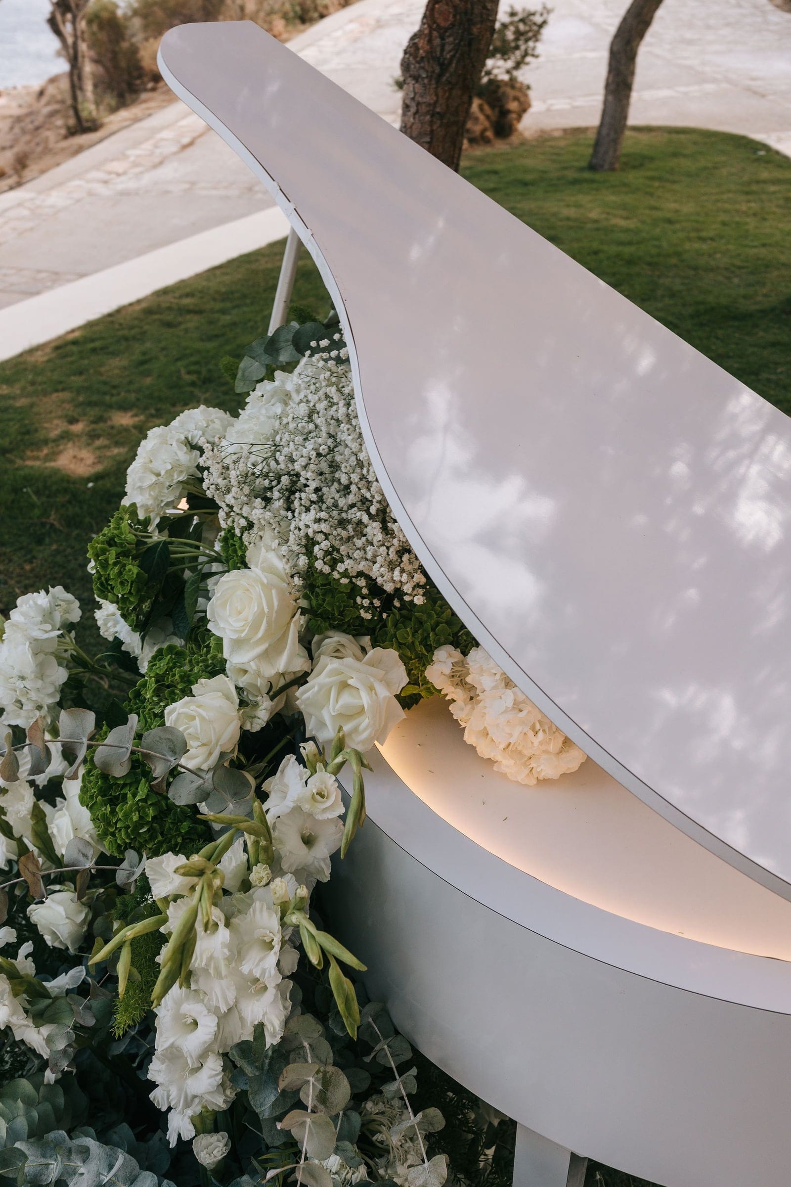 A white grand piano on a lawn at Cycladic Gem Luxury Villa on Ios Island, Santorini, Greece, its curved lid and glowing underside framed by a lush arrangement of white garden roses, white hydrangeas, baby's breath, and silvery eucalyptus. Green hydrangea heads and white gladioli add depth and texture to the wedding flowers spilling along its base. Soft daylight falls across the glossy white lid, with a stone path and an olive tree in the blurred background.