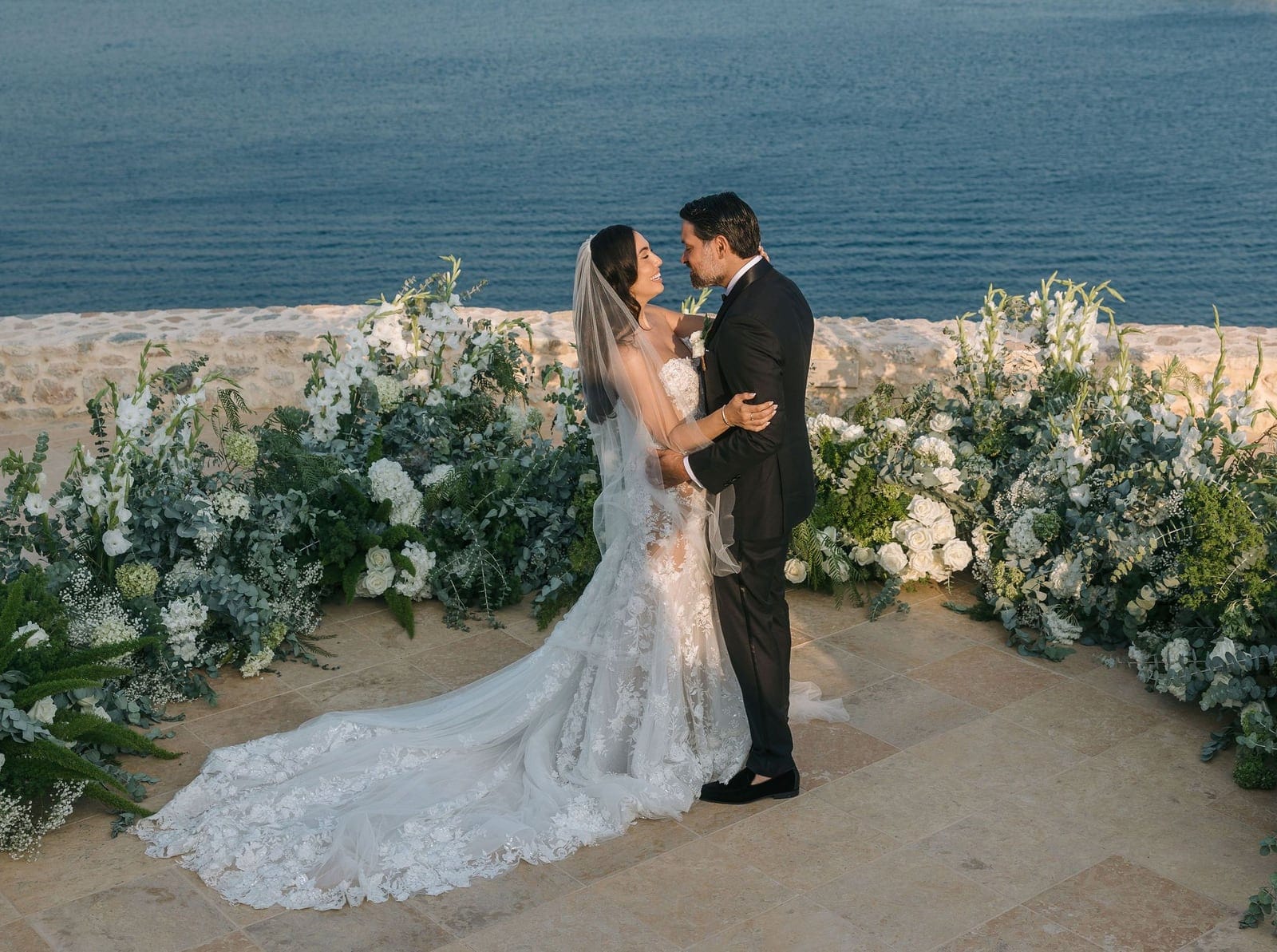 A bride in a lace gown with a long train and a flowing veil shares a close, smiling moment with her groom in a dark suit, standing on a stone terrace before a low wall, with the deep blue Aegean sea stretching behind them. Lush arrangements of white roses, gladioli, hydrangeas and silvery eucalyptus frame the couple, and the warm evening light softens the scene. These are wedding flowers at the Cycladic Gem Luxury Villa on Ios, in the Santorini area of Greece.