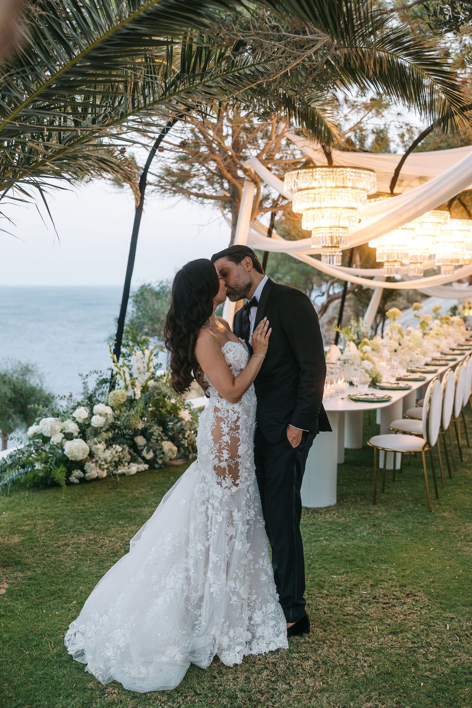 A bride in a lace fitted gown with a sheer lace bodice and long train kisses her groom in a black tuxedo under a canopy of white draped fabric and tiered crystal chandeliers, with a palm frond overhead. Beside them, white hydrangeas and white garden roses spill across the grass, and a long reception table with gold-framed chairs stretches away toward the sea. This is a wedding flower and styling scene at Cycladic Gem Luxury Villa on Ios Island, Santorini, Greece.