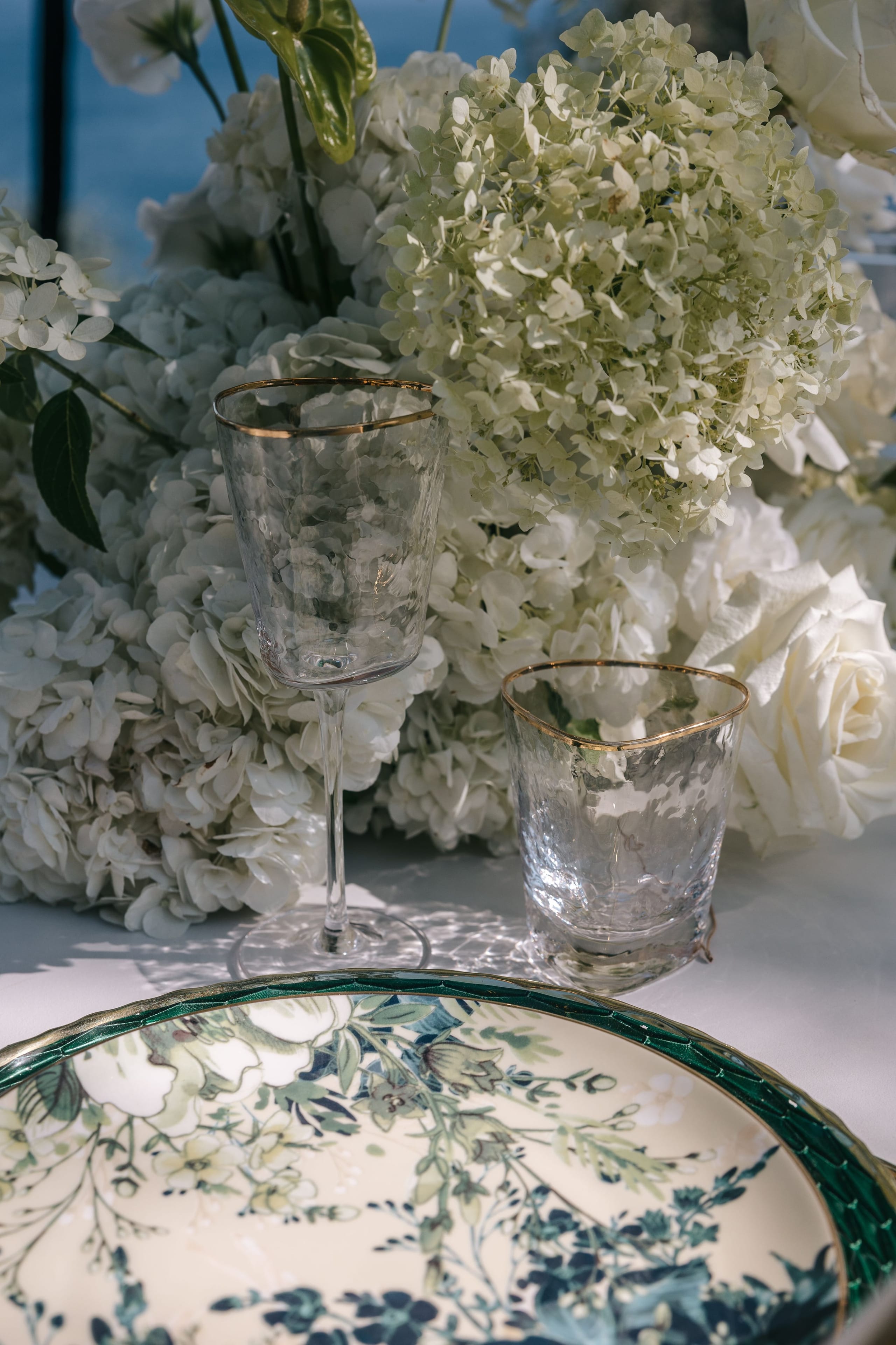 Cream and white hydrangeas, with a few white garden roses, fill the background of this wedding table setting at Cycladic Gem Luxury Villa on Ios, Santorini, Greece. In the foreground, two hand-hammered glass goblets with gold rims stand on a white cloth, and a green-rimmed plate painted with delicate blue and cream florals sits below them. Soft shadows and a blue sea glimpsed behind the flowers give it a calm, sunlit feel.