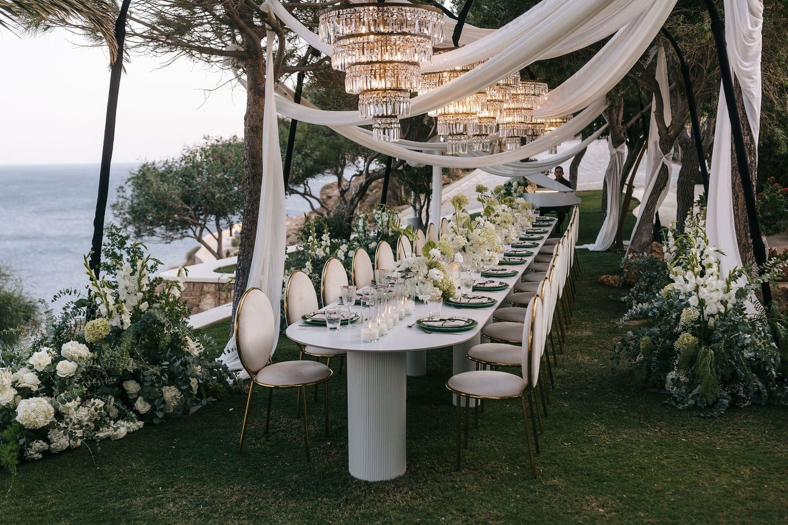 A long, oval white wedding banquet table set on a grassy clifftop under draped white fabric and tiered crystal chandeliers, with the sea glimpsed through the pines beside it. White garden roses, hydrangeas and gladioli spill in lush arrangements along both sides of the table, with candles and green-rimmed plates set for guests seated on cream velvet chairs with gold legs. This wedding table setting in Ios, Santorini, Greece, at Cycladic Gem Luxury Villa, has a soft, romantic evening glow.