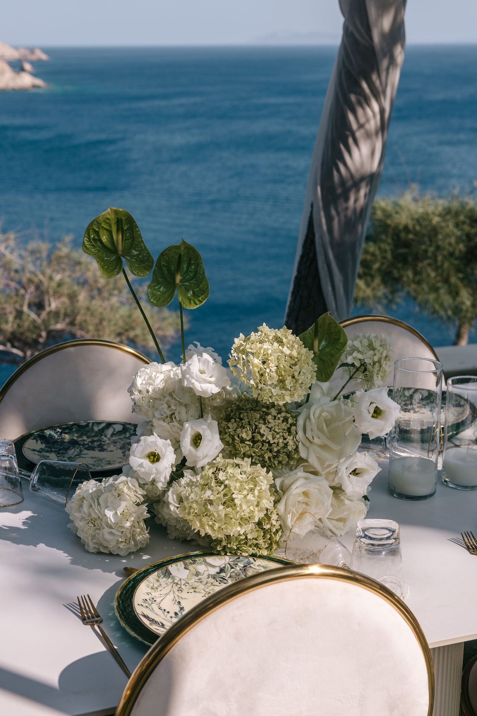 A wedding table centrepiece of white lisianthus, cream garden roses and pale green hydrangeas, with two tall anthurium stems rising above it, set on a white table at Cycladic Gem Luxury Villa on Ios island, Santorini, Greece. Gold-rimmed cream chairs and patterned plates frame it, and beyond the terrace the deep blue Aegean sea stretches to the horizon under bright sunlight.