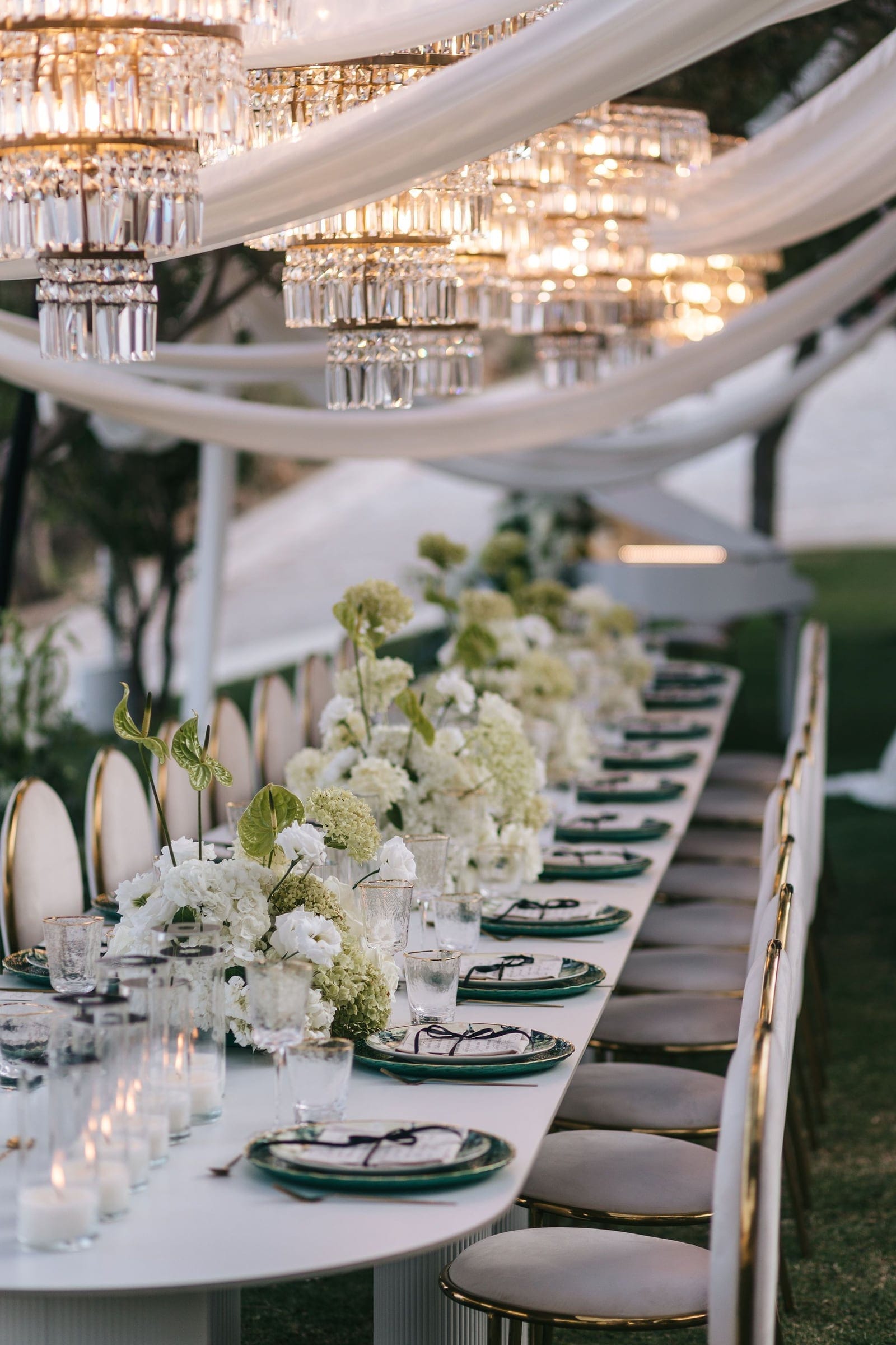 A long white banqueting table stretches into the distance beneath a canopy of draped white fabric and tiered crystal chandeliers, their warm lights glowing. Along it run low arrangements of white hydrangeas, white blooms and green anthurium, with green-rimmed plates, folded place cards tied with dark ribbon, and pale velvet chairs with gold frames. This is a wedding table setting at Cycladic Gem Luxury Villa on Ios Island, Santorini, Greece.