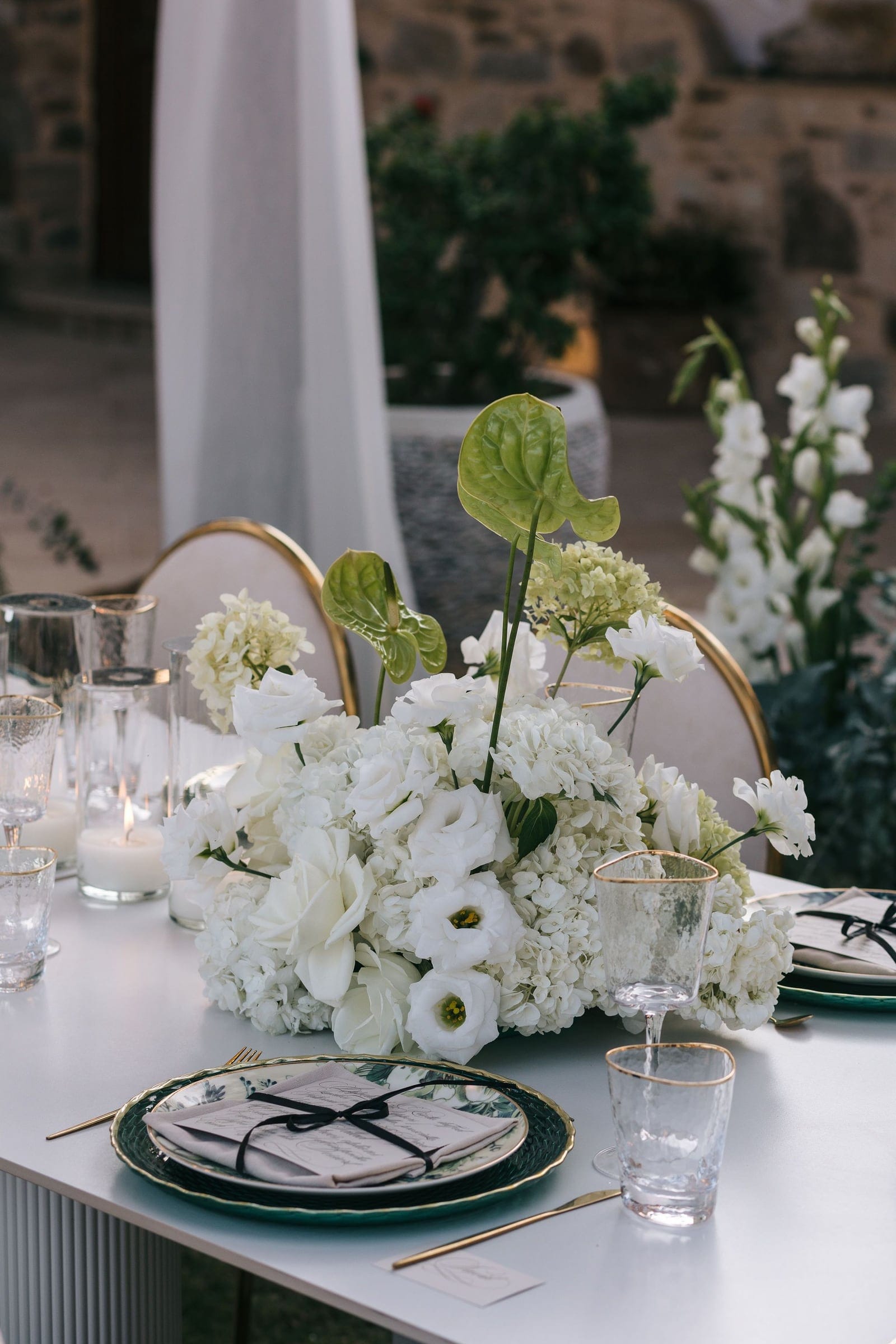 A low, lush centrepiece of white hydrangeas, lisianthus and white garden roses, with tall green anthurium leaves rising from the middle, sits on a white table at Cycladic Gem Luxury Villa on Ios Island, Santorini, Greece. Its wedding table flowers are set among gold-rimmed glasses, a pair of white candles in glass hurricanes, and dark green plates tied with black ribbon. Behind it, white gladioli stand in a tall arrangement against the warm stone wall of a courtyard in the soft evening light.