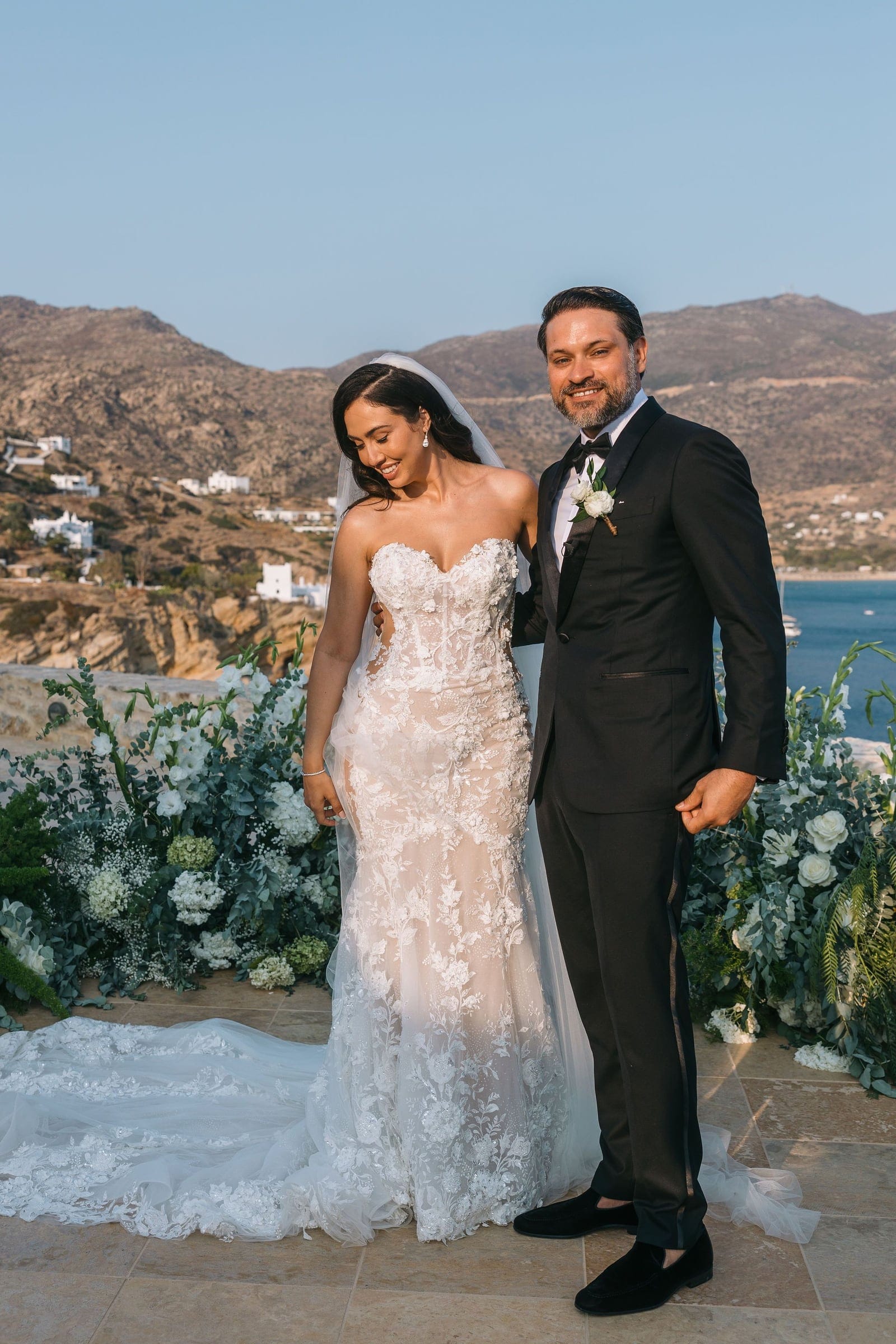 A bride in a strapless, fitted lace gown with a long chapel train and a sheer veil stands beside a smiling groom in a black tuxedo and bow tie, a white rose boutonniere on his lapel. Behind them, a low bank of white roses, hydrangeas and eucalyptus lines a stone terrace at the Cycladic Gem Luxury Villa on Ios, Santorini, Greece, with the sea and white-washed houses on the sunlit hillside beyond. These wedding flowers are arranged in soft, airy greens and whites.