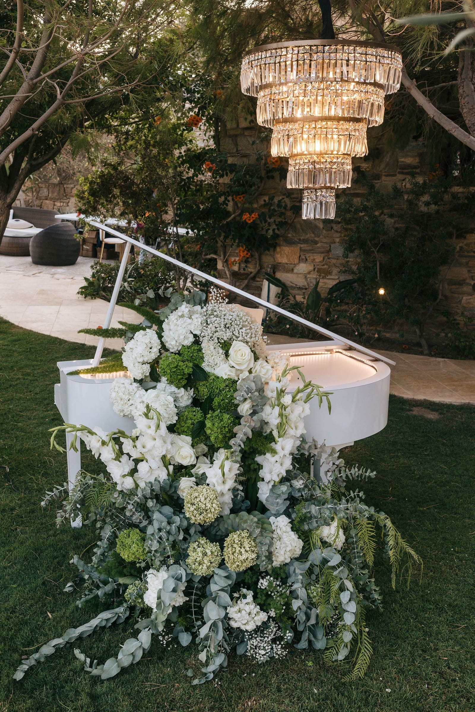 A white grand piano on a lawn at Cycladic Gem Luxury Villa on Ios island, Santorini, Greece, is smothered in a lush wedding floral arrangement of white roses, white gladioli, green hydrangea heads, baby's breath and trailing silver eucalyptus. Above it hangs a tiered crystal chandelier, its lights glowing against a warm stone wall and dusky garden trees.