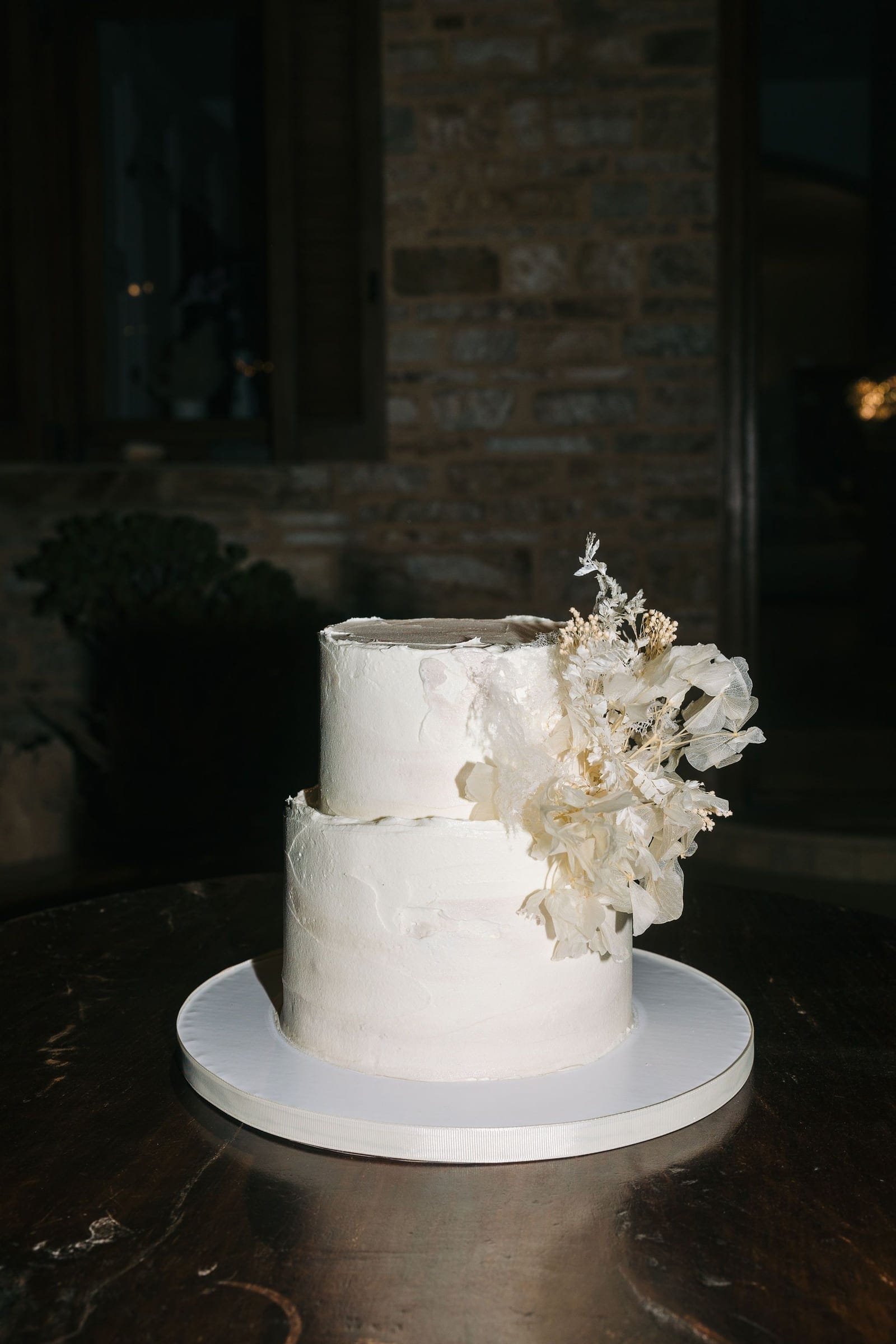 A two-tier wedding cake with smooth, softly textured white icing, set on a white board on a dark wooden table. Pale dried hydrangea blooms, delicate white sprays and small dried seed heads cascade down one side of the upper tier, adding a soft, rustic touch. Behind it, a brick wall and warm light create a quiet, intimate mood at this wedding at Cycladic Gem Luxury Villa on Ios Island, Santorini, Greece.