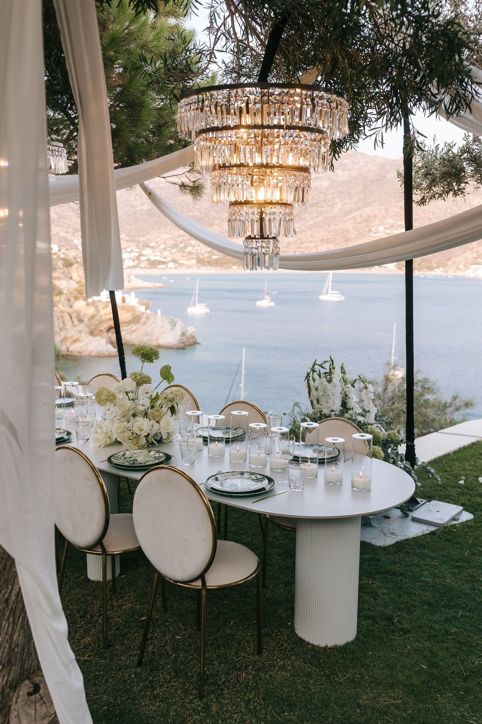 A long white wedding dining table sits on lawn beneath draped white fabric and a tiered crystal chandelier, with the blue bay of Ios Island, Greece, stretching behind it and sailboats at anchor. Low arrangements of white blooms, likely roses and hydrangea, flank the table, which is set with green-rimmed plates, clear glasses and pillar candles on the sheer, airy terrace of Cycladic Gem Luxury Villa.