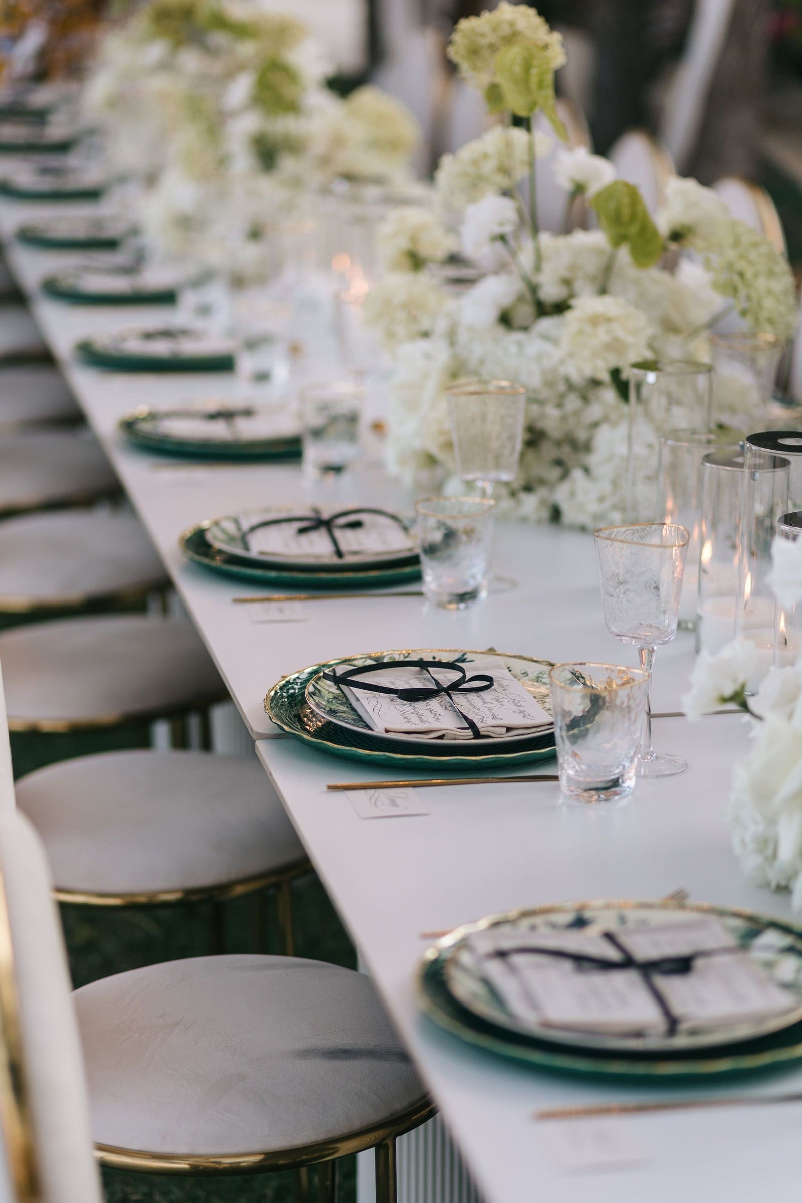 A long white banquet table runs diagonally into the distance, dressed with white and pale green hydrangea centrepieces, their full heads softened by a few green blooms. Each place setting has green-rimmed plates topped with a cream menu tied in a black ribbon, flanked by clear etched glassware and gold cutlery, with pale upholstered gold-legged chairs along one side. These wedding table flowers were arranged at Cycladic Gem Luxury Villa on Ios Island, Santorini, Greece, in soft, golden evening light.