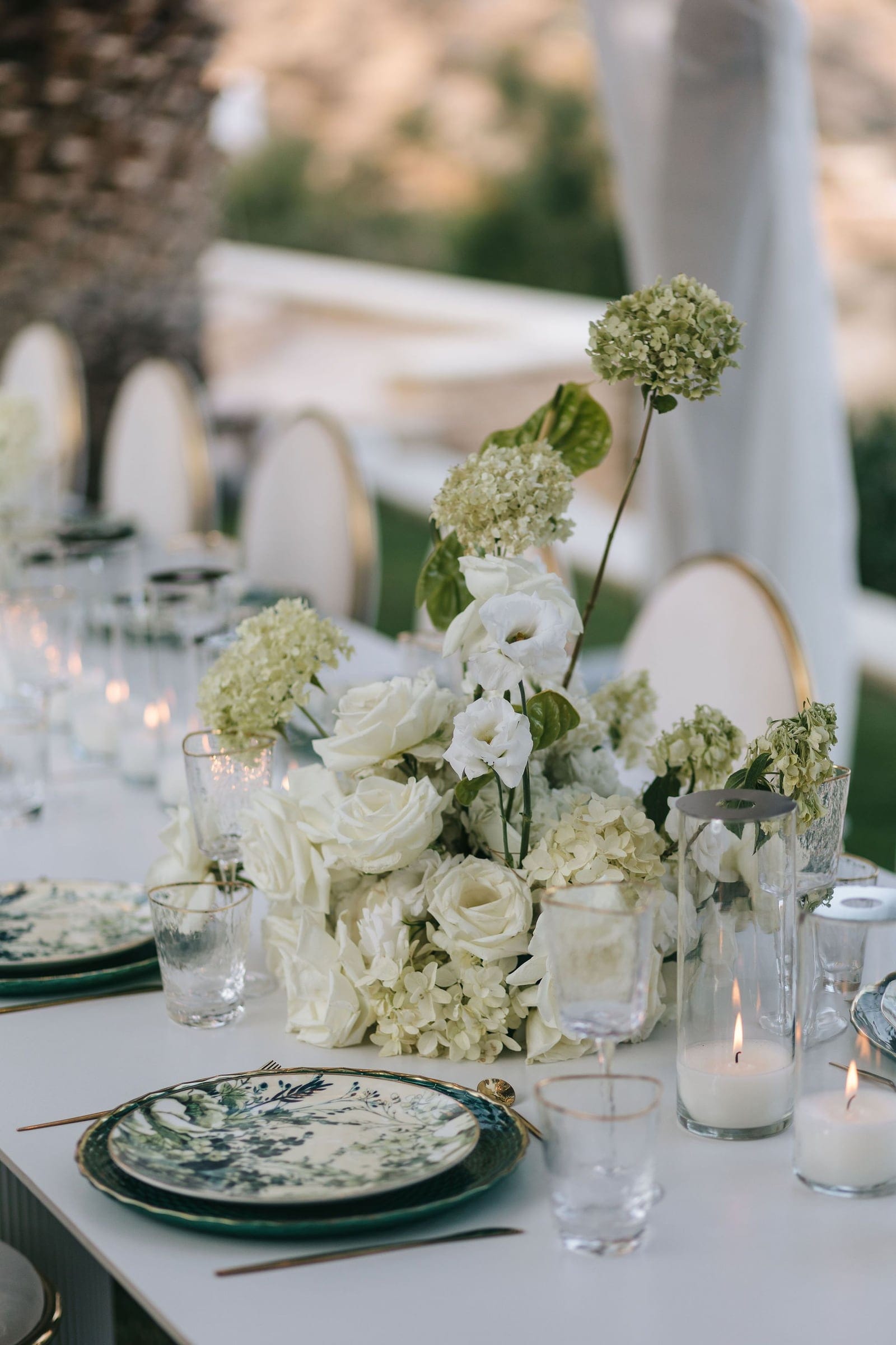 A low centrepiece of white garden roses, white lisianthus and pale green hydrangeas, with tall single hydrangea stems rising from it, sits on a long white table set with green-rimmed botanical plates and clear glassware. Tall pillar candles glow beside it in the soft evening light of a wedding reception at Cycladic Gem Luxury Villa on Ios Island, Santorini, Greece.