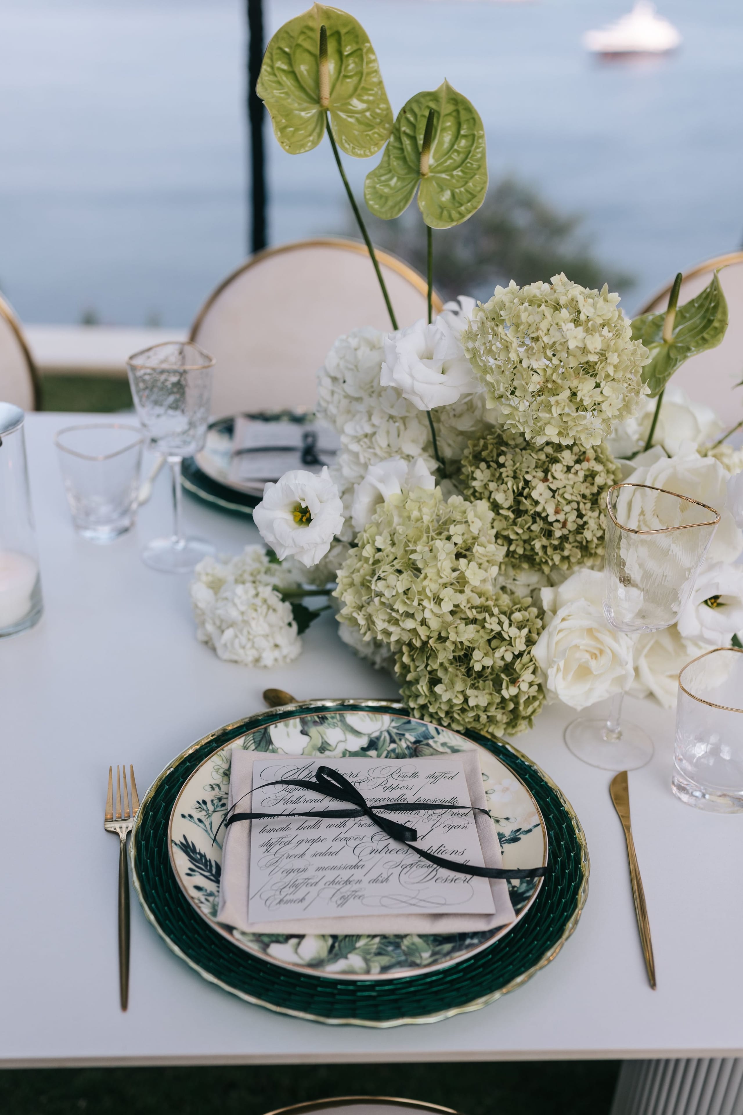 Green hydrangeas and white lisianthus fill a low centrepiece on a white outdoor wedding table, with green anthurium blooms rising above it. In the foreground, a dark green and gold charger holds a floral-patterned plate, a menu card tied with a black ribbon and gold cutlery. Soft sea and a distant boat fill the background, set at the Cycladic Gem Luxury Villa on Ios, Santorini, Greece.
