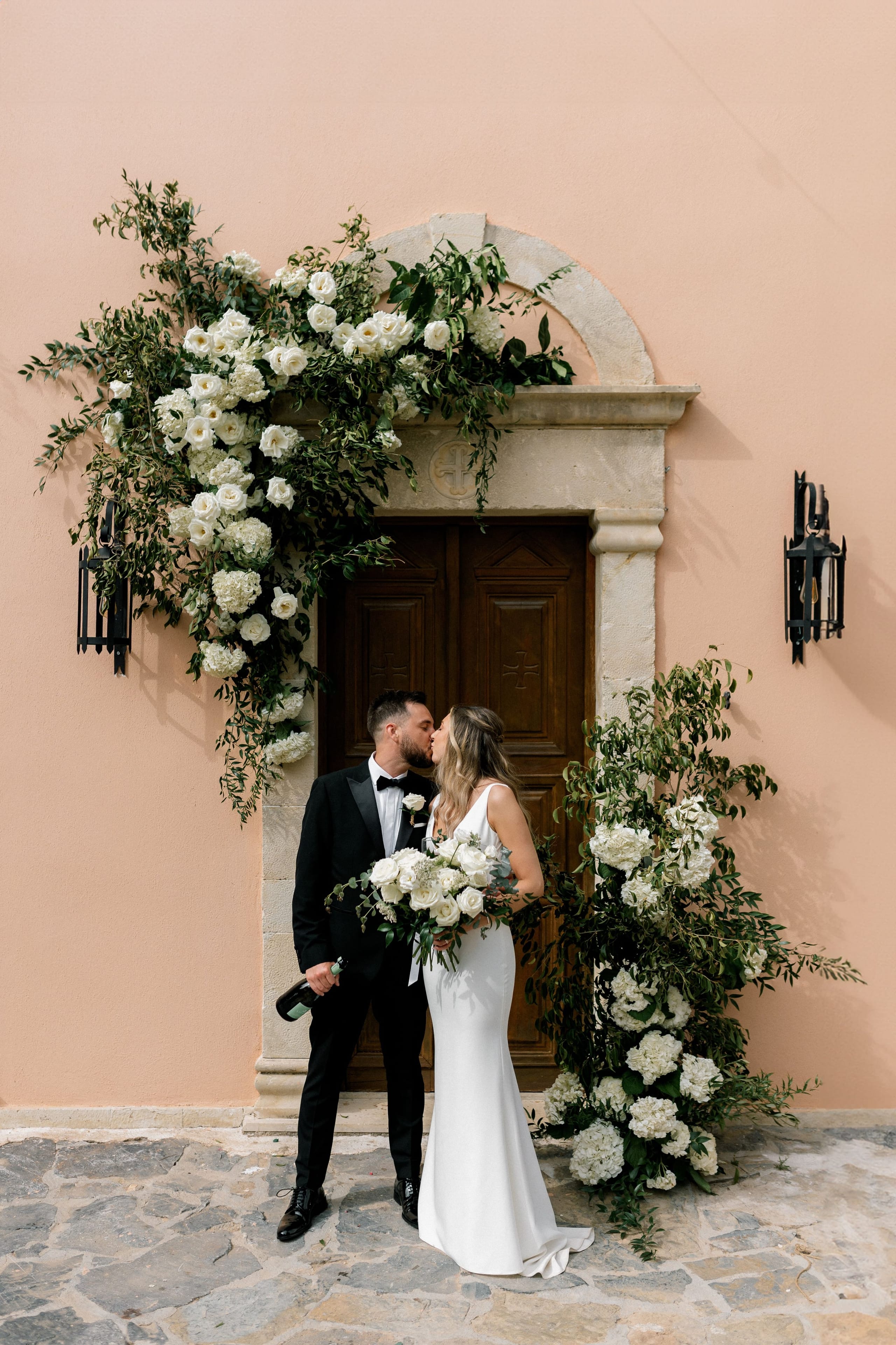 A bride in a fitted white satin gown and a groom in a black tuxedo kiss in front of a dark wooden door set in a pale stone archway on a peach-coloured wall. Trailing over the arch and down the left side is a lush installation of white roses, white hydrangeas and green foliage, with a matching cascade on the right, and the bride holds a white rose and hydrangea bouquet. This wedding flower arrangement is at AgrecoFarms in Adele, Crete, Greece.