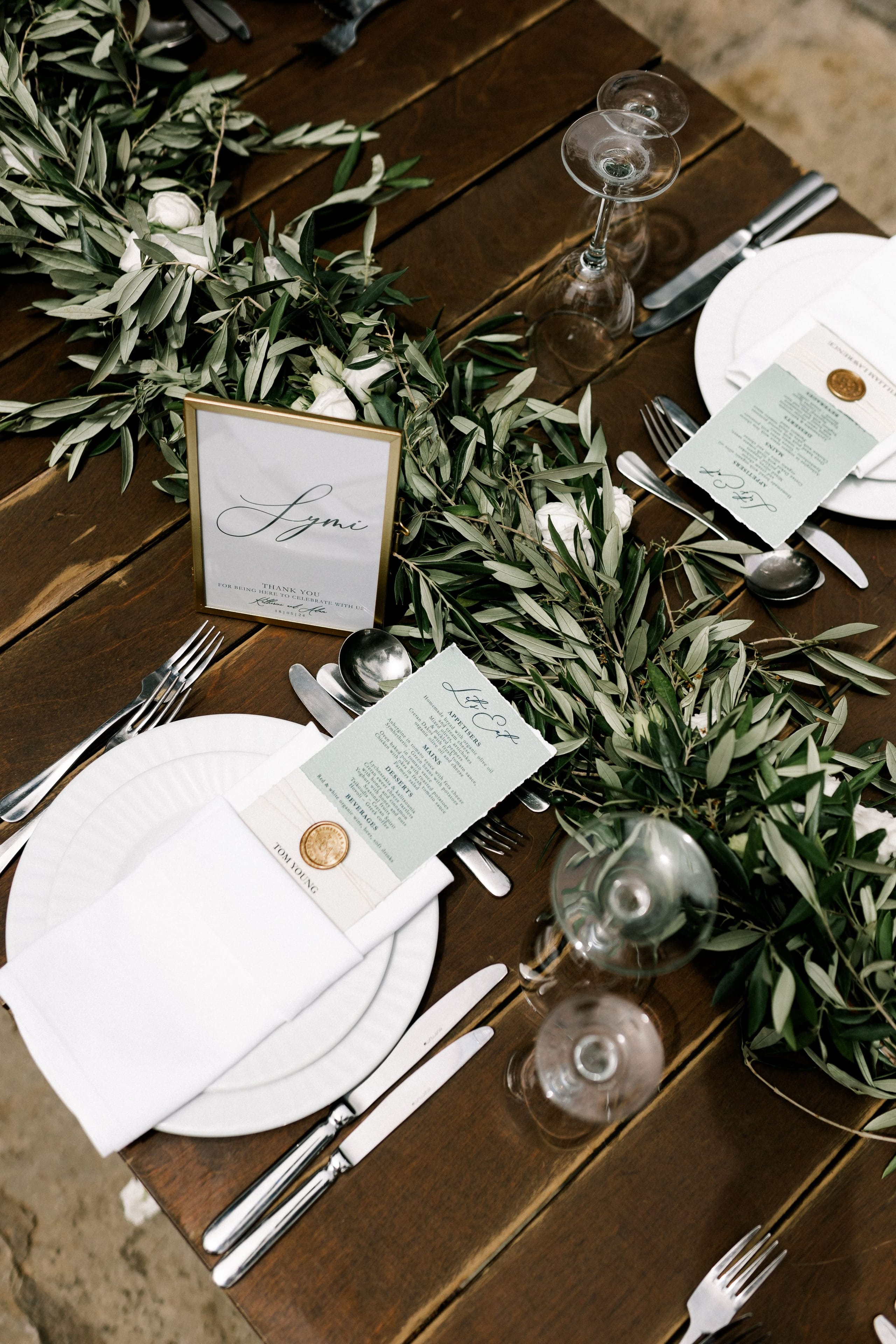 A rustic wooden wedding table dressed with a long runner of olive branches threaded with small white roses, set in Crete, Greece. Each place setting has a white plate with a folded white napkin, polished silver cutlery, and clear wine glasses, and a sage-green menu card sealed with a gold wax stamp. A gold-framed table name card reading "Lysmi" stands among the greenery.