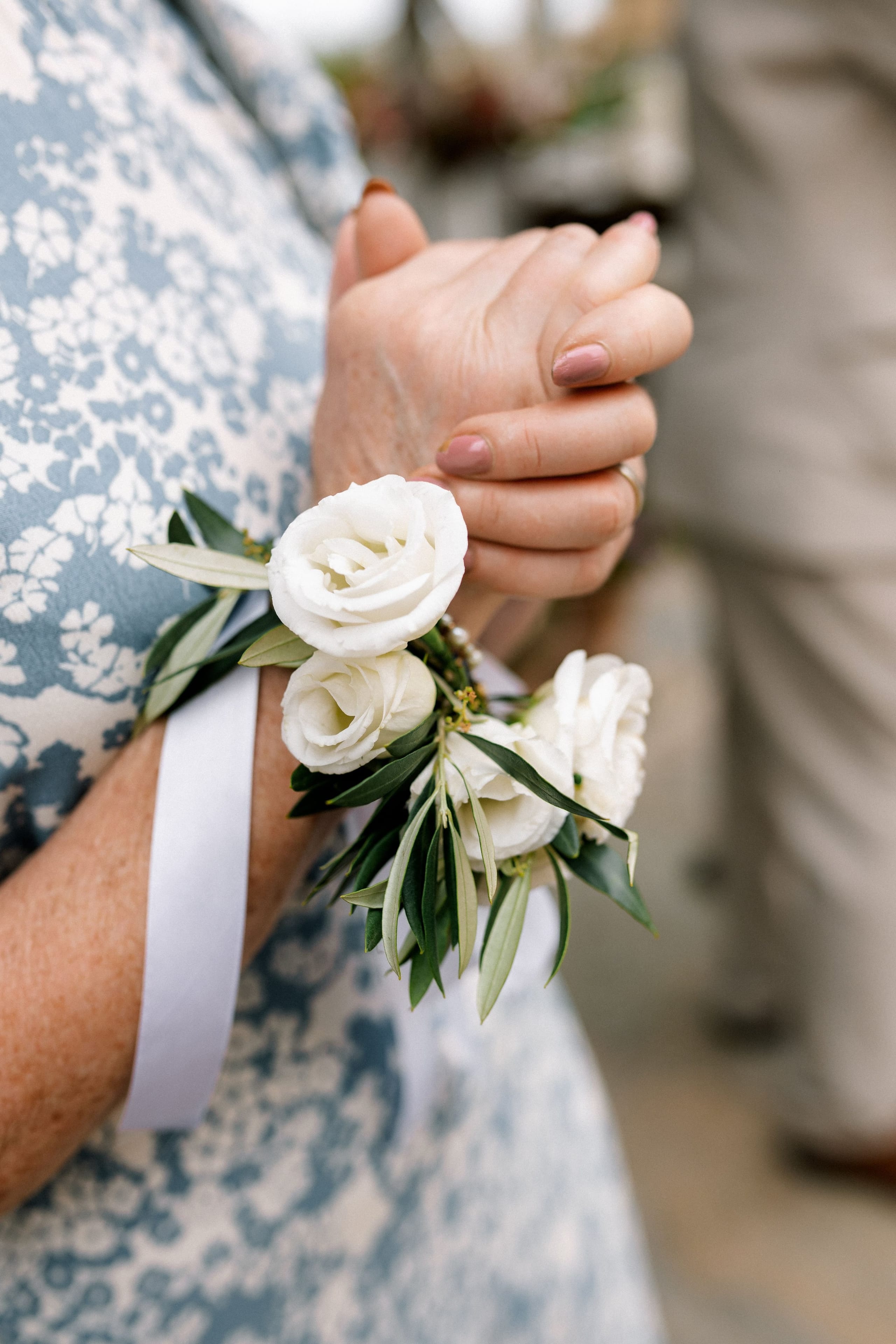 A close view of a white wrist corsage of small white roses with slender olive leaves, tied with a pale ribbon around the wrist of a woman in a blue and white floral dress. Her hand rests gently over another hand at her waist, and the soft background suggests an outdoor wedding flower detail at AgrecoFarms in Adele, Crete, Greece.