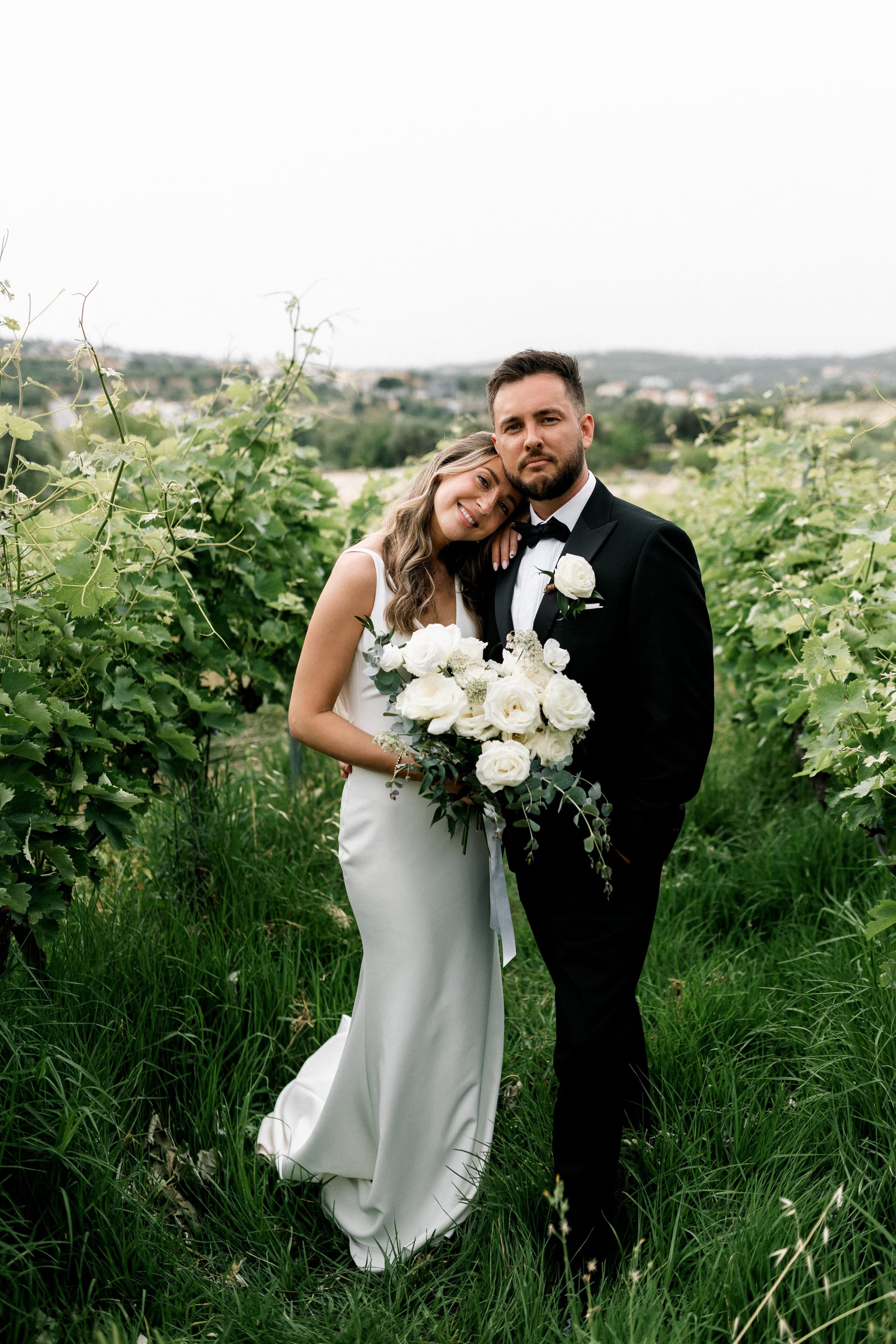 A bride in a fitted white satin gown holds a cascading bouquet of white garden roses, white hydrangea florets and trailing eucalyptus, while she leans her head against the shoulder of a groom in a black tuxedo with a white rose boutonniere. They stand together between green vine rows in tall grass, with soft hills and a pale sky behind them in this wedding photo from AgrecoFarms in Adele, Crete, Greece.